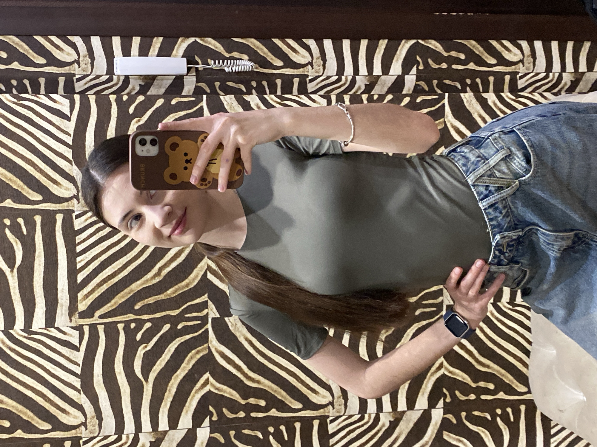 A young woman is taking a selfie in Votkinsk, Russia. She is lying on a bed with a zebra-print blanket. She is wearing a green t-shirt and blue jeans. She is holding a smartphone with a bear-shaped case. She has long brown hair. There is a white telephone attached to the headboard.