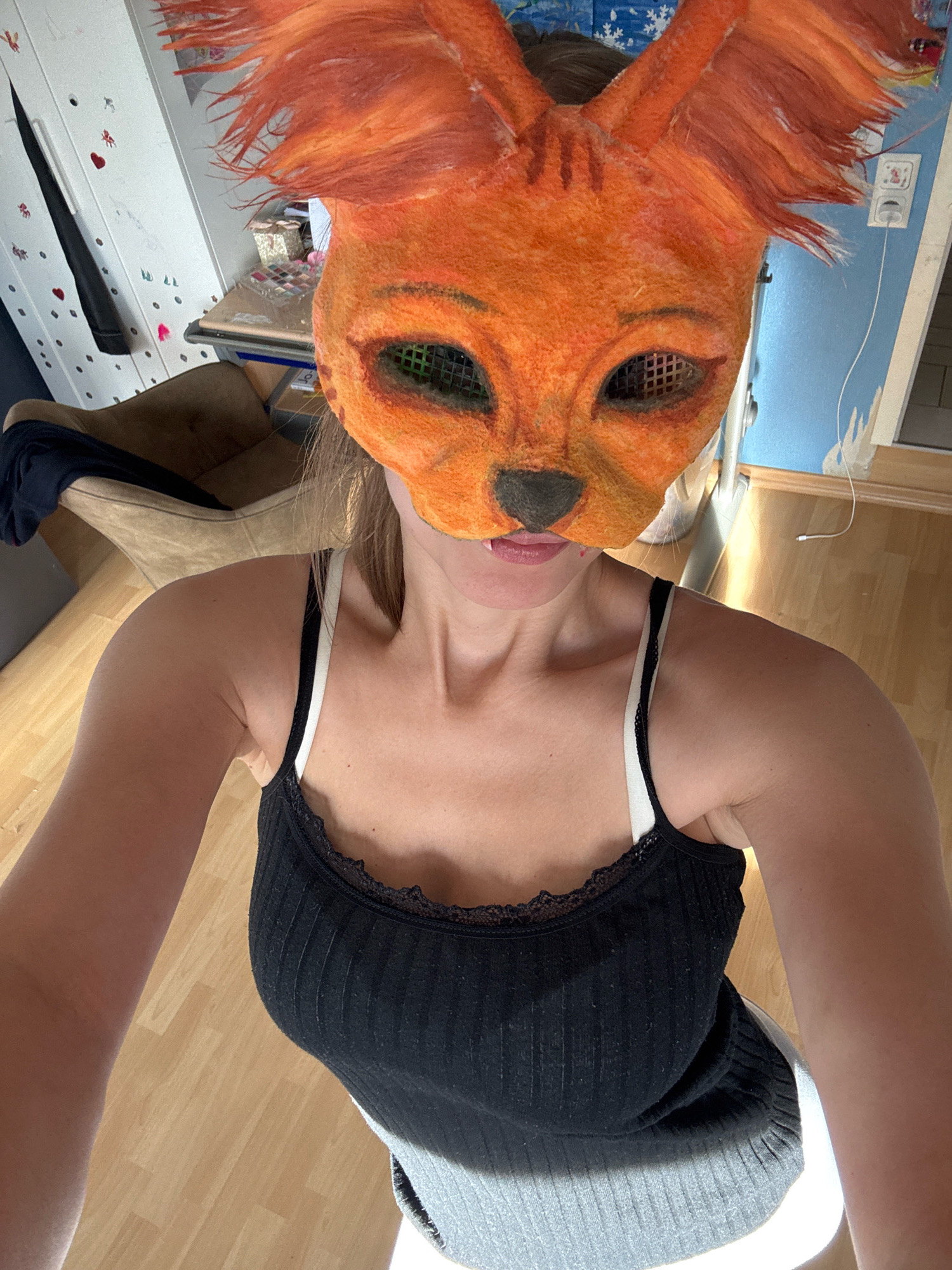 In a residential indoor setting, a young woman, seen from the chest up, poses for a photograph. She wears a handmade fox mask covering her face, characterized by large ears and orange fur. The mask has black mesh over the eye openings and a black nose. She is also wearing a black tank top with white straps, adorned with black lace trim. The background reveals a light brown wooden floor and a portion of a wall painted light blue. To the left, part of a brown armchair is visible, and further left, a white closet door is adorned with small red and pink stickers. The overall impression is that of a casual, personal photograph taken within a home environment in Überherrn, Germany.