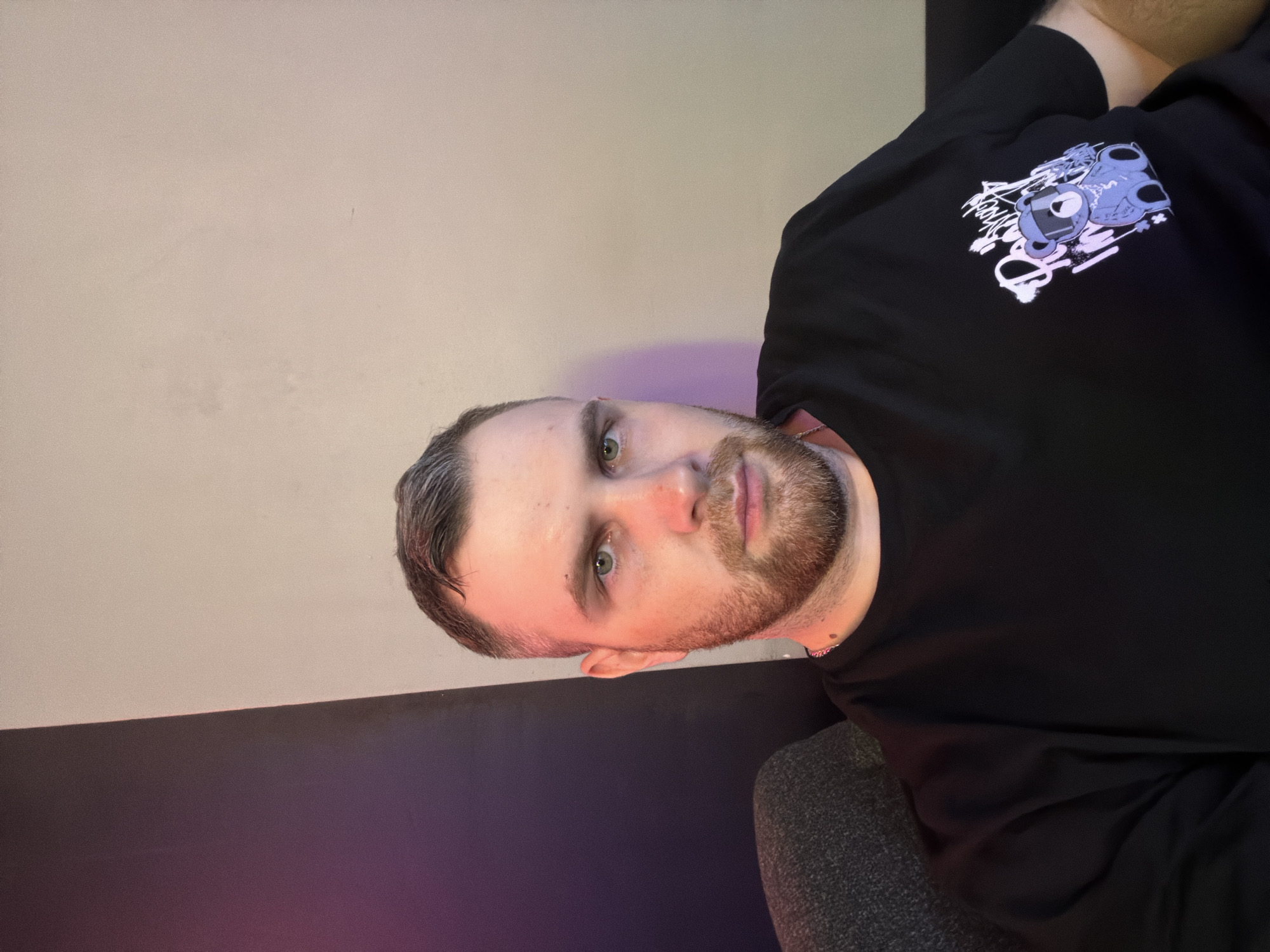 A medium shot of a man with short brown and gray hair, light eyes, and a short beard. He is wearing a black long-sleeved shirt with a graphic design on the upper left. The background consists of a beige upper wall and a lower section that transitions from dark gray to purple. The scene is indoors and appears to be a personal space. There is no information to indicate that the picture was taken in Izhevsk, Russia.