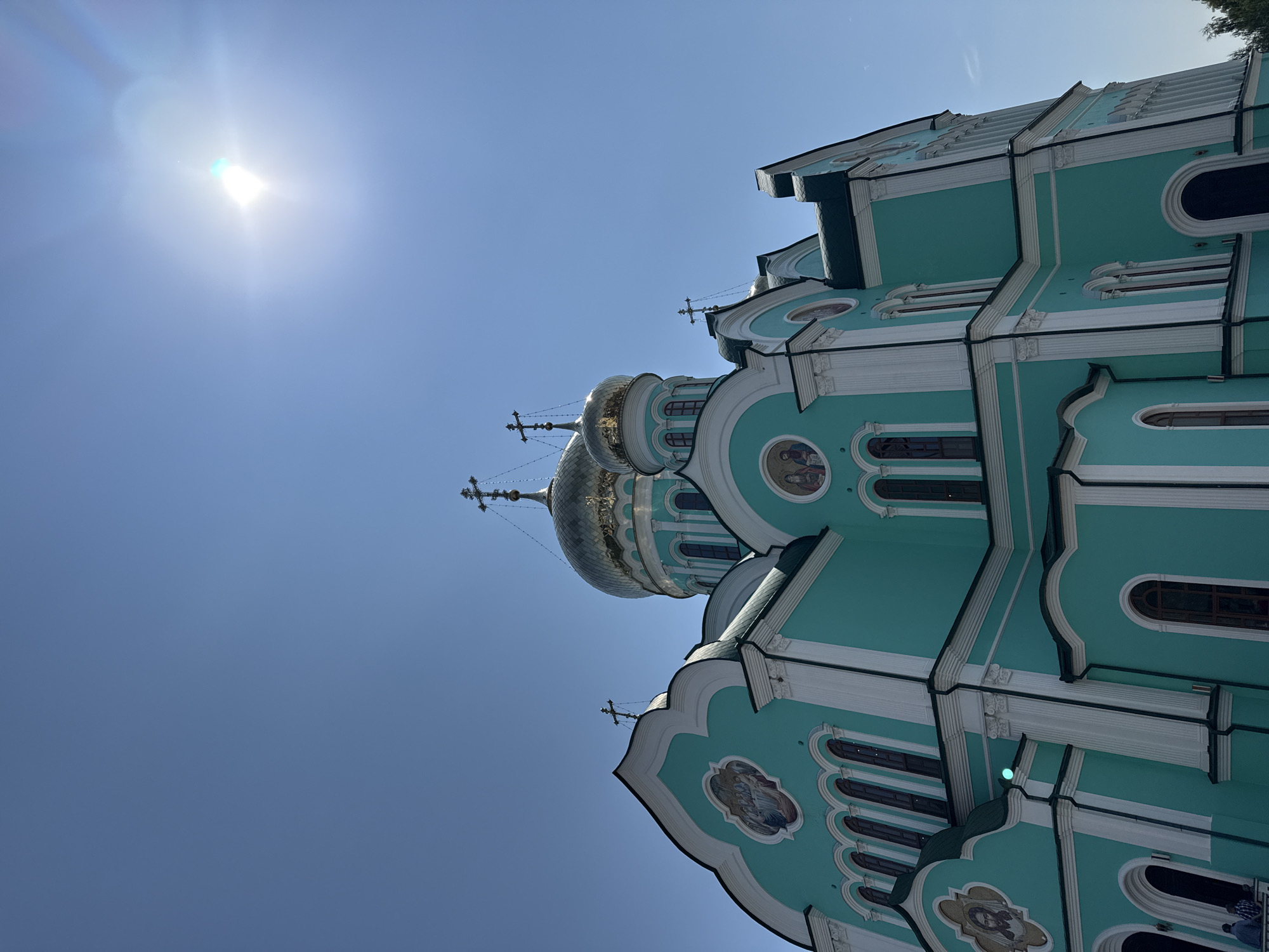 A low-angle, full-shot captures the exterior of a turquoise Orthodox church against a clear, bright blue sky. The sun is high in the sky and to the right, creating a lens flare. The church has multiple golden domes topped with crosses. The building's walls are adorned with white trim, arched windows, and circular mosaics depicting religious figures. Several crosses are visible on the roofline, and two figures are standing on a balcony in the lower left corner. The church appears to be a prominent landmark in the city.