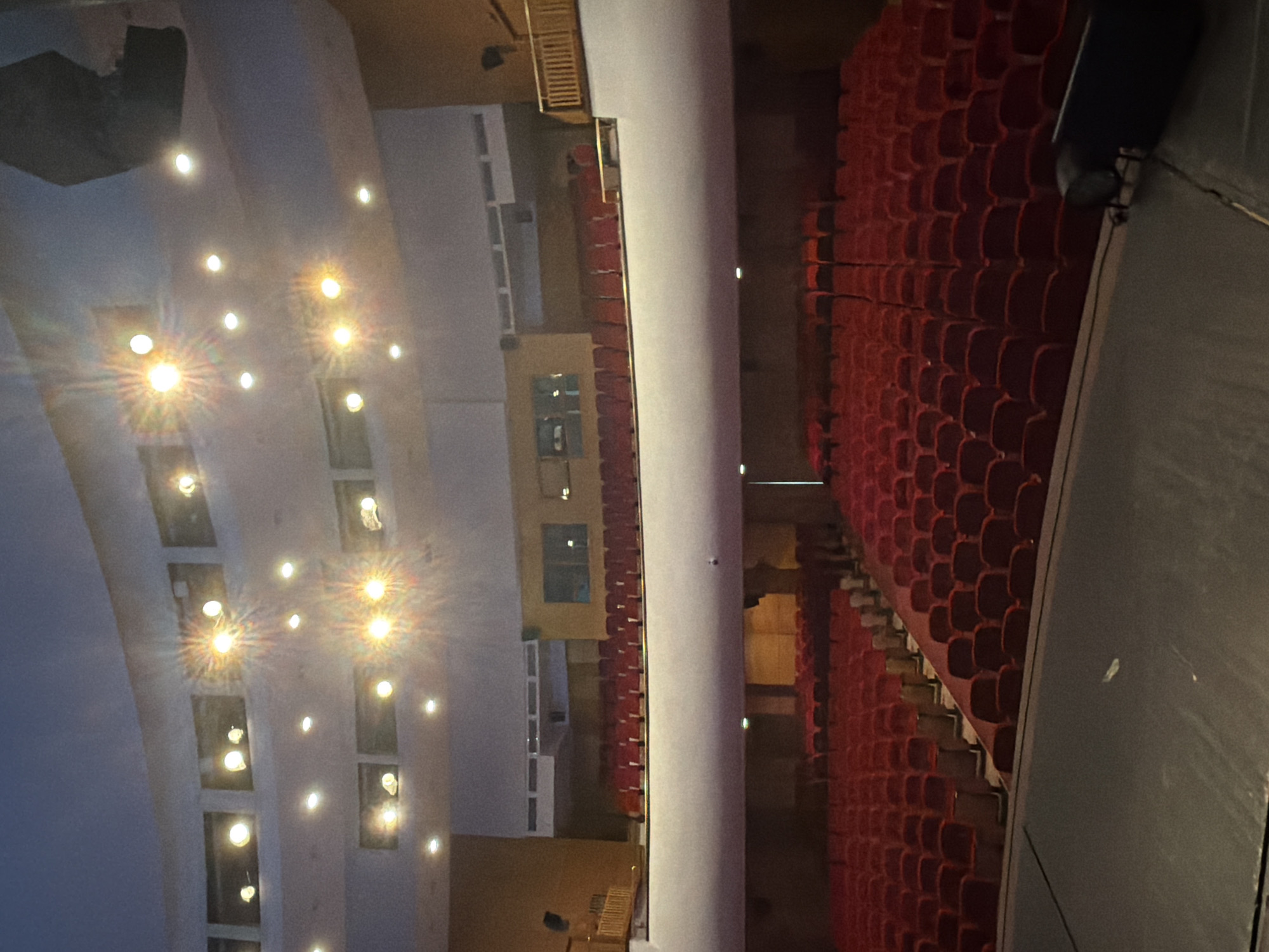 The image depicts the interior of the Ancient Theatre of Philippopolis in Plovdiv, Bulgaria. The view is oriented from the stage towards the audience seating area. Red upholstered seats are arranged in tiers, facing the stage. A central aisle separates the seating sections. The upper level of the theatre is visible, with additional seating. The ceiling is arched and illuminated by numerous spotlights. The lighting creates a bright atmosphere, contrasted with shadows in the seating area. No people are visible in the frame. A sound system component is visible stage right.