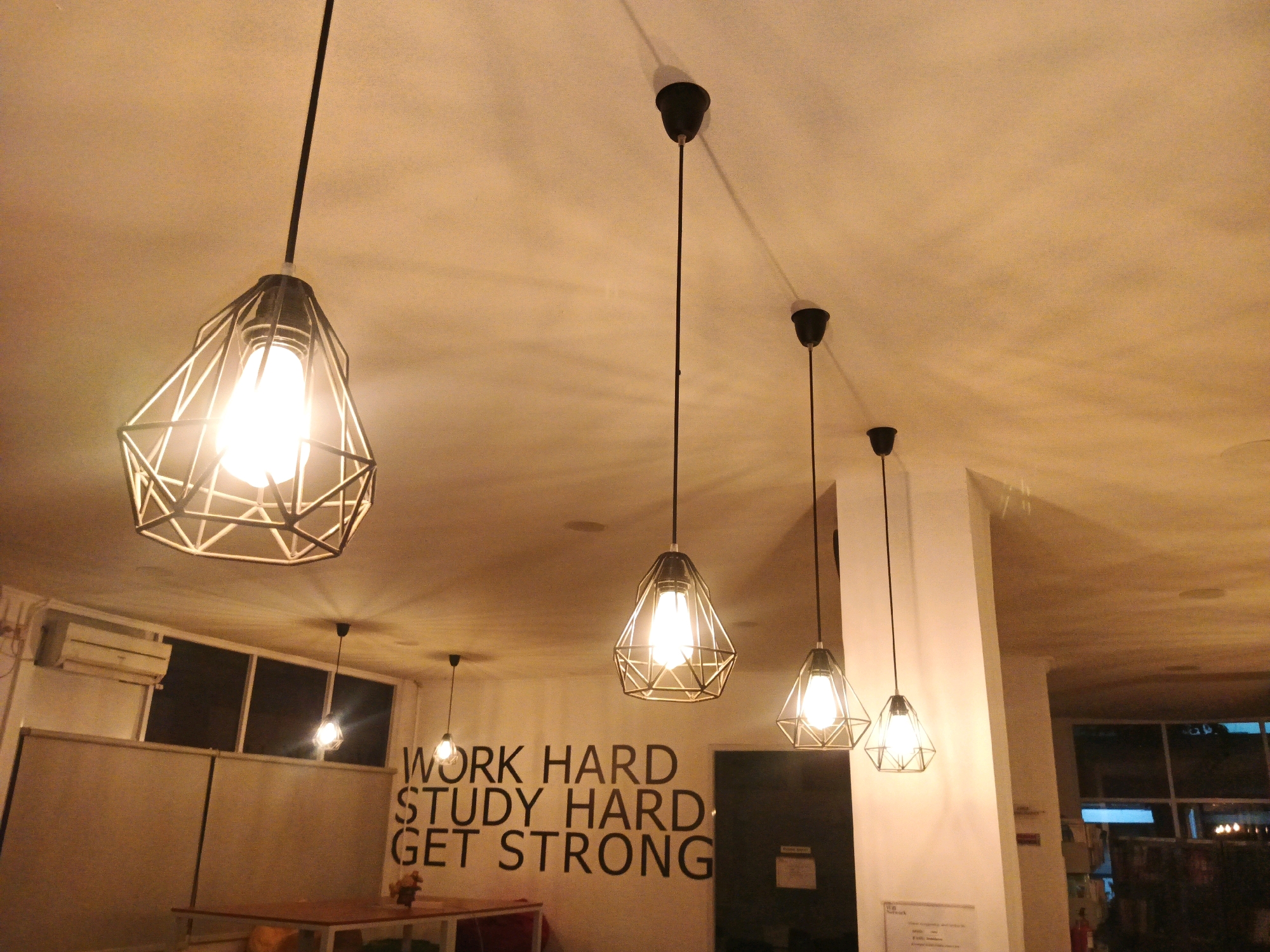 The image shows the interior of a room with several pendant lights hanging from the ceiling. The lights have geometric, cage-like shades and are illuminated. In the background, there is a wall with text that reads "WORK HARD STUDY HARD GET STRONG". To the left, there are windows and an air conditioning unit. A table and chairs are visible in the foreground. The scene appears to be a study or common area. There is no specific information or visual cue to determine the exact location in Bandung City, Indonesia.