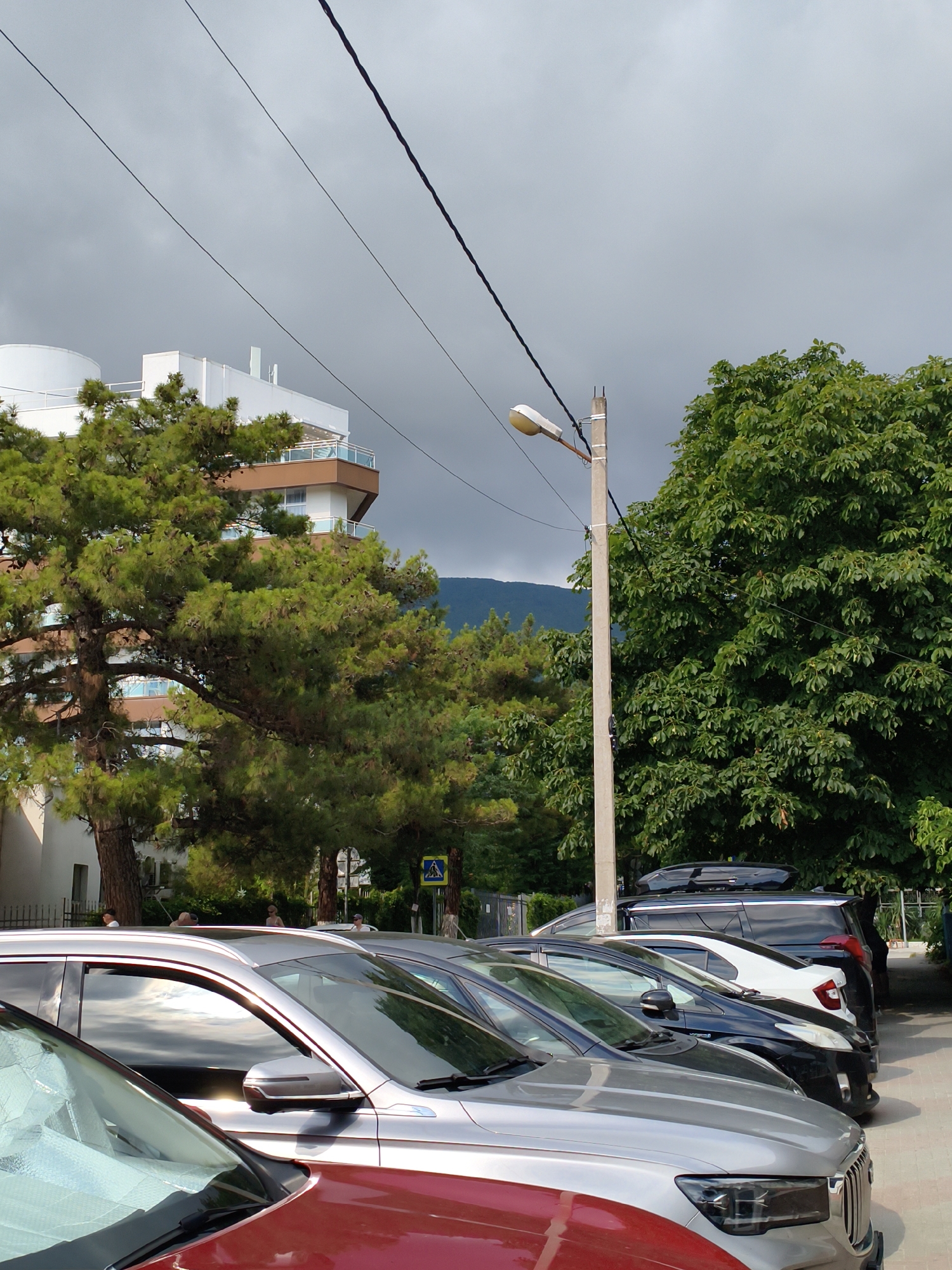 The image shows a parking lot in Gelendzhik, Russia, with several cars parked in a row. In the background, there is a modern multi-story building with balconies and a white circular structure on top. Tall green trees, including pine trees, are visible on the left and right sides of the image, with lush foliage. The sky is overcast with gray clouds. A concrete lamppost with a street light is positioned to the right of center. A pedestrian crossing sign is visible behind some trees. People are sparsely visible in the background, some standing near the building and others possibly walking. The cars are parked on an asphalt surface. The overall scene depicts a typical urban outdoor environment in Gelendzhik.