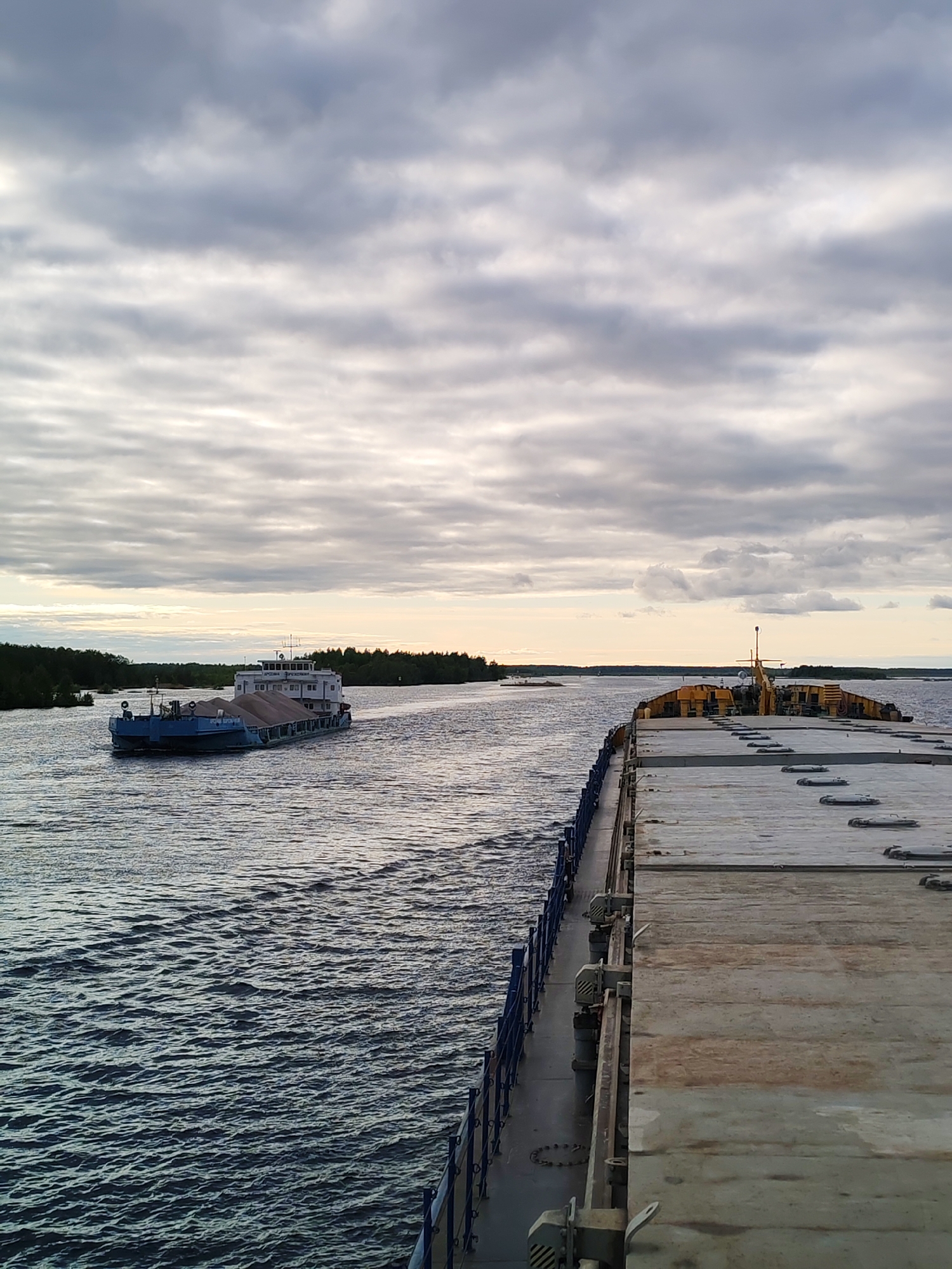 The image shows a wide body of water, likely a river or a wide channel, under an overcast sky. In the distance, a blue tugboat pulls a barge loaded with gray material, possibly gravel or sand. The barge is moving towards the left side of the frame. The vantage point is from another large barge or vessel, visible in the foreground. A blue railing runs along the left side of this barge. The deck of the foreground barge is made of light gray, weathered material. The shoreline is visible in the distance with a dark green line of trees. The lighting suggests either early morning or late afternoon. There is no evidence in the image to suggest that it is located in the city of Bereg, Russia.