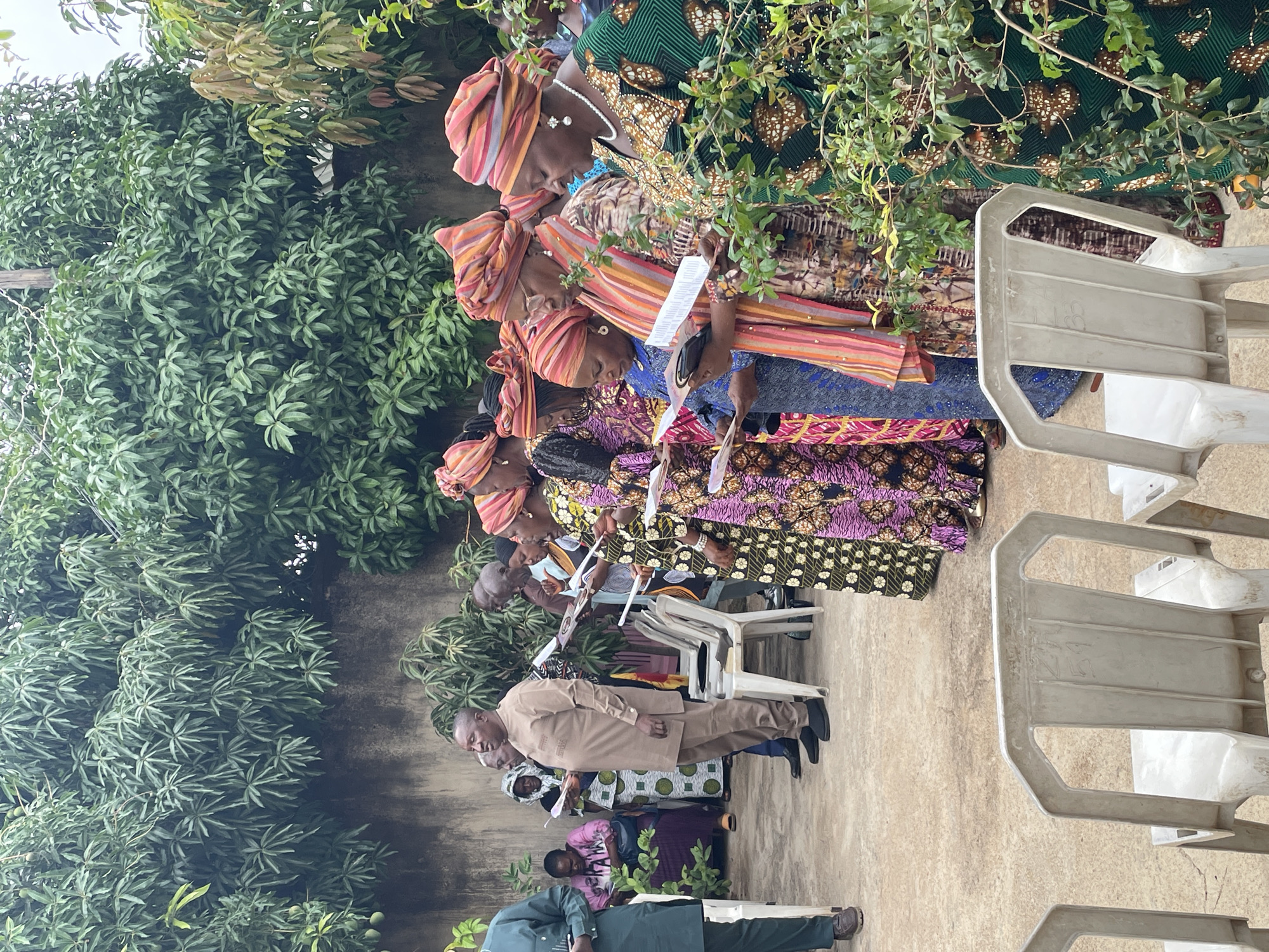 This image captures an outdoor gathering in Jos, Nigeria, featuring a group of people engaged in a communal activity. The setting is verdant, with dense green foliage and trees providing a natural backdrop and some overhead cover, indicating a tropical or subtropical environment. The ground appears to be a light-colored paved or concrete surface.
The subjects are primarily adults, predominantly women, dressed in vibrant, patterned traditional Nigerian attire, including colorful head wraps (gele) and flowing dresses or skirts. Several men are also present, some in similar traditional outfits, while others wear more Western-style clothing, such as a man in a light brown suit standing prominently in the foreground.
The activity involves people standing and some seated on white plastic chairs. Many individuals are holding white papers, seemingly reading from them or following along with a program, suggesting an organized event like a church service, a memorial, or a community meeting. Their expressions are generally focused and attentive, conveying a sense of quiet engagement.
Notable details include the time of day, which appears to be daylight, likely morning or afternoon, under natural, diffused lighting, possibly indicating an overcast sky or shaded area. The chairs are simple plastic designs, some stacked, others in use, suggesting a practical, informal outdoor venue. While papers are held, any specific text on them is not legible. One white plastic chair has faint markings, possibly "B2". The overall atmosphere is one of collective participation in a respectful gathering amidst nature.