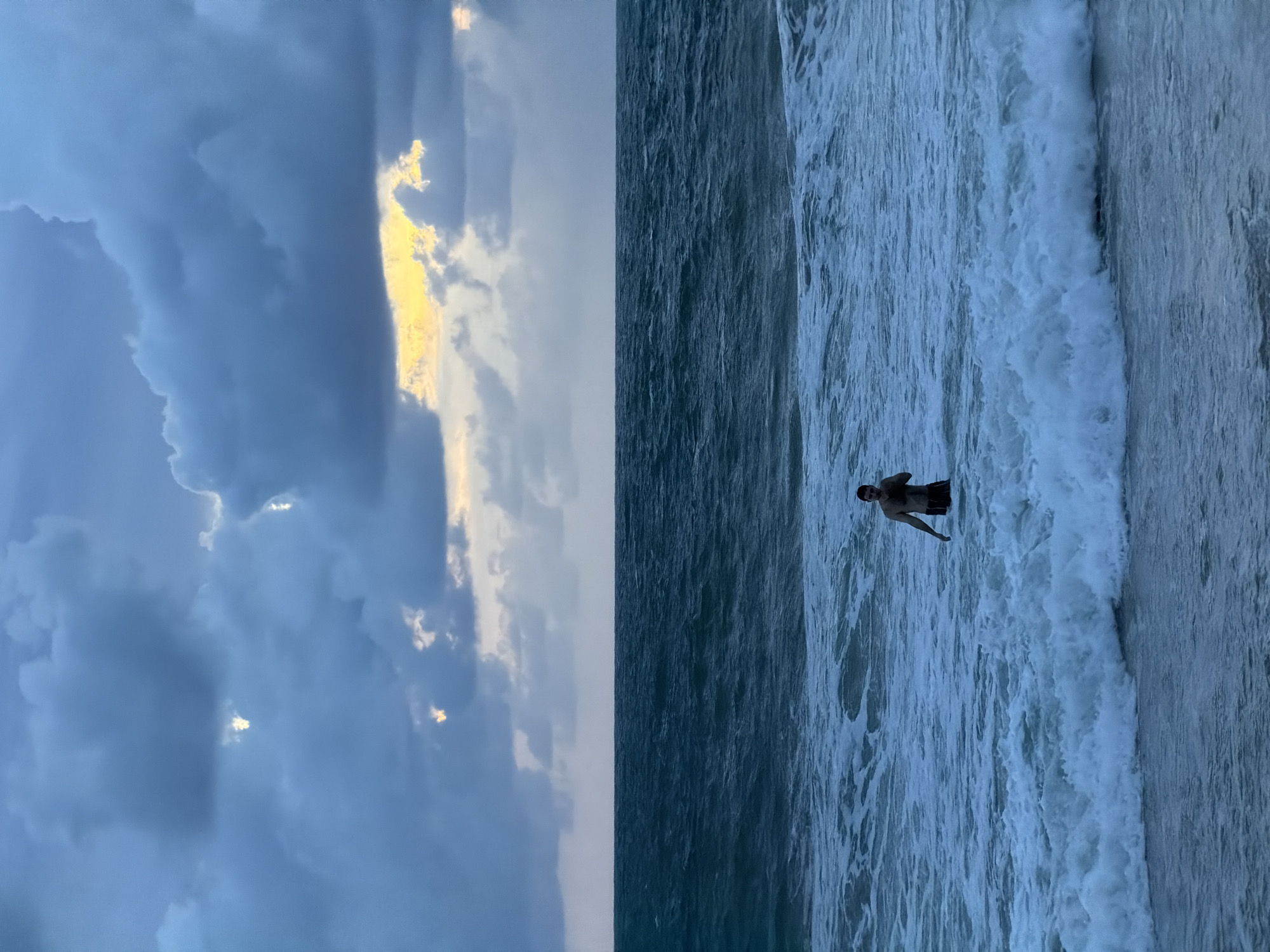 A vertical outdoor shot shows a person waist-deep in the surf at Choeng Thale, Thailand. The person is near the shore. The turbulent ocean is a mix of dark and light blues, with a frothy wave rolling toward the person. The left side of the frame shows a dramatic sky with heavy, dark clouds. Hints of light break through the clouds near the horizon.