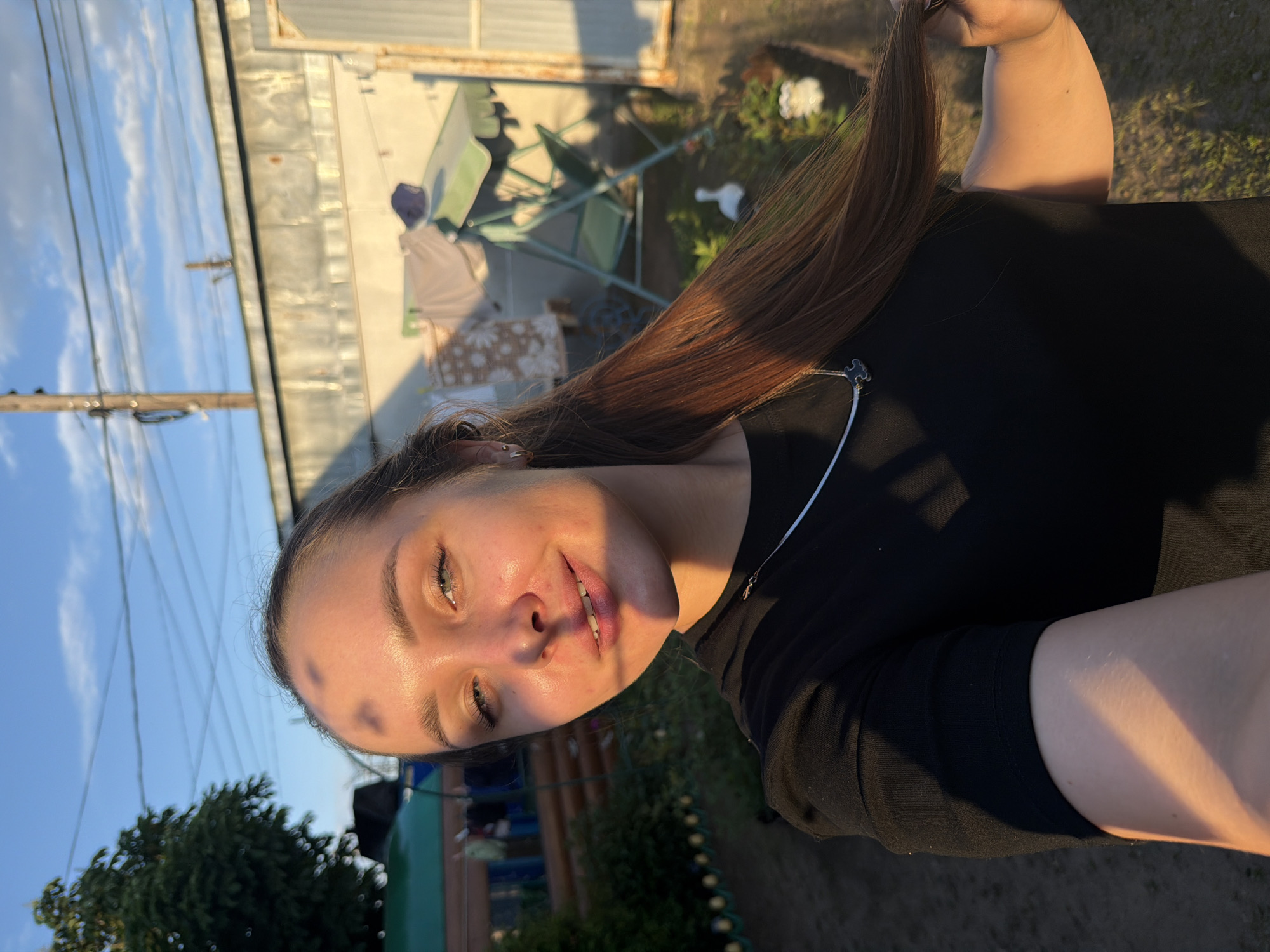 A young woman with long brown hair and blue eyes is taking a selfie outdoors. She is wearing a black t-shirt and a silver necklace with a dark pendant. Her face is partially illuminated by the setting sun, casting a warm glow and some shadow patterns on her forehead and cheek. She is smiling slightly and holding a lock of her hair in her right hand. The background appears to be a backyard in a rural or suburban setting, possibly in Chaikovsky, Russia, given the presence of a wooden structure, a green swing set with a canopy, clothes hanging on a line, and greenery. The sky is blue with some white clouds and power lines are visible.