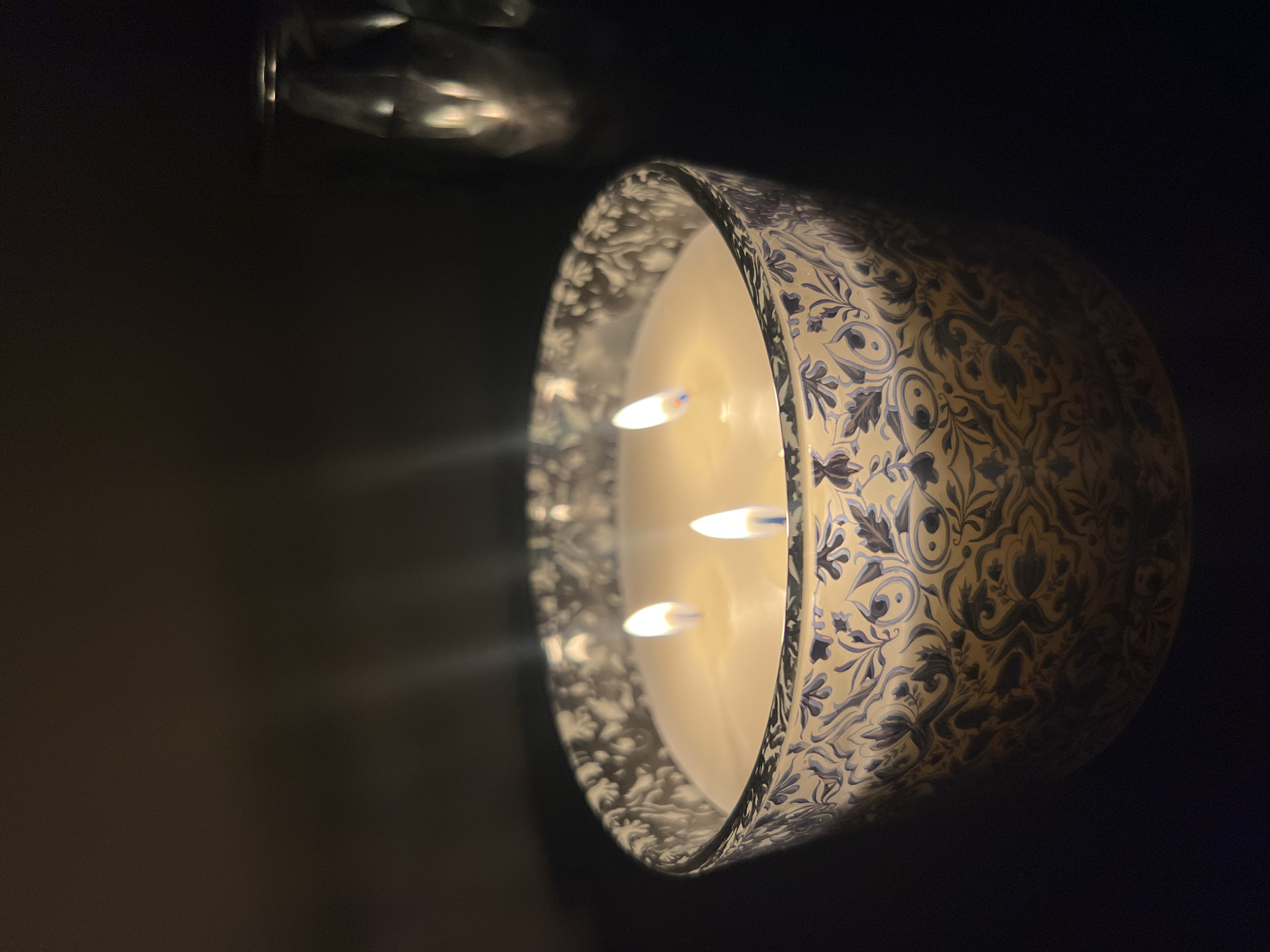 A lit candle with three wicks rests in a patterned glass container. The candle has a cream-colored wax. The glass container is decorated with a floral pattern in shades of blue and off-white. The background is dark, with some indistinct reflective shapes in the upper left corner. There is no evidence to indicate a specific location in Sofia, Bulgaria or any other location. There are no people present in the image.