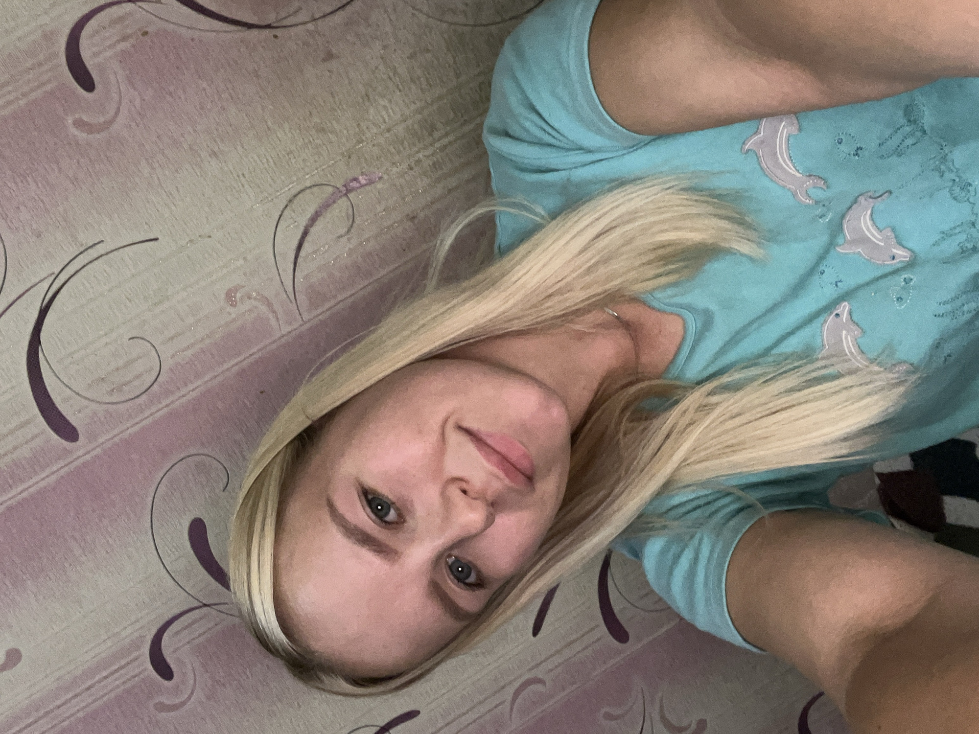 A young Caucasian woman with blonde hair and blue eyes is captured in a selfie. She is wearing a turquoise t-shirt with a dolphin print and a thin silver necklace. The background consists of wallpaper with a striped pattern and purple swirls. The lighting suggests an indoor environment.