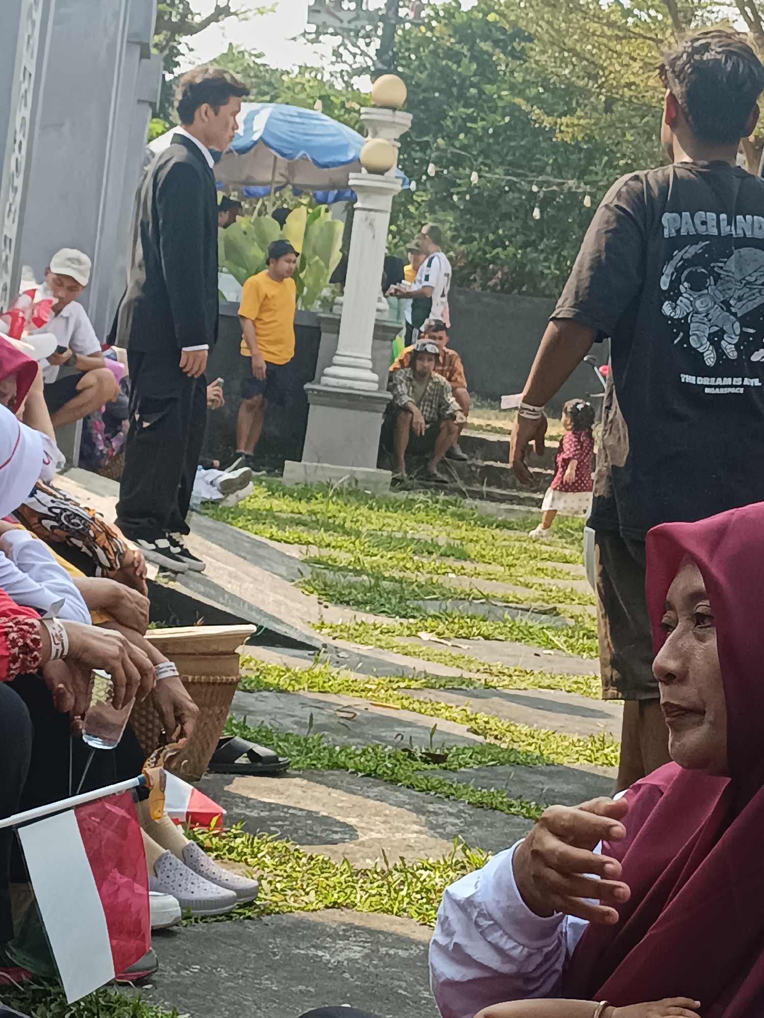 The image depicts an outdoor gathering, possibly in Sleman, Indonesia, given the Indonesian flag prominently displayed. In the foreground, several people are seated. A woman wearing a maroon hijab and a white shirt is visible on the right, looking to the side. To her left, another person's hands are visible, one holding a glass of water, with an Indonesian flag planted nearby. Several individuals are seated in the background, some in casual attire, while a man in a black suit stands in the center. The environment features lush green grass, paved pathways, and trees in the background, suggesting a park or an outdoor event venue. The lighting indicates it is daytime, likely sunny.