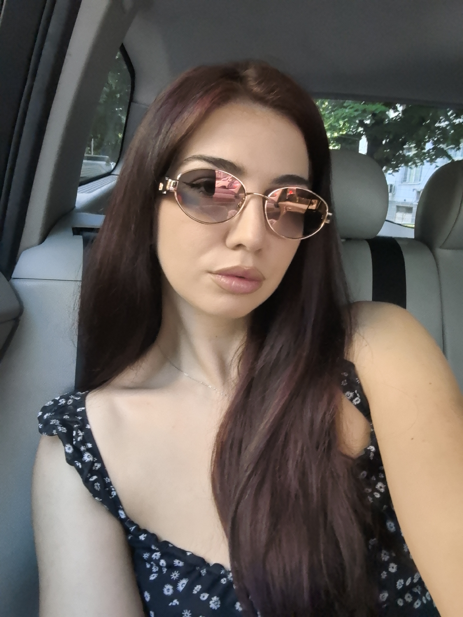 A young woman is captured in a close-up selfie, seated in the passenger side of a car during what appears to be a bright day. She has long, straight dark brown hair, possibly with reddish highlights, that frames her face and flows over her shoulders. She wears stylish oval sunglasses with gold frames and rose-tinted lenses, which reflect elements of the car's interior and the bright outdoor environment, including buildings and trees. Her lips are slightly parted in a subtle pout, appearing full and glossy. She is wearing a dark-colored top or dress with a small, white floral or daisy pattern.
The setting is clearly the interior of a vehicle, with glimpses of grey car seats featuring black accents and a dark grey door panel. Through the car windows, vibrant green foliage from trees and parts of light-colored buildings are visible, suggesting an urban or suburban street scene typical of a place like Stara Zagora, Bulgaria. The bright lighting conditions indicate a sunny day. The woman is looking directly at the camera, conveying a composed and casual mood. There is no visible text in the image.