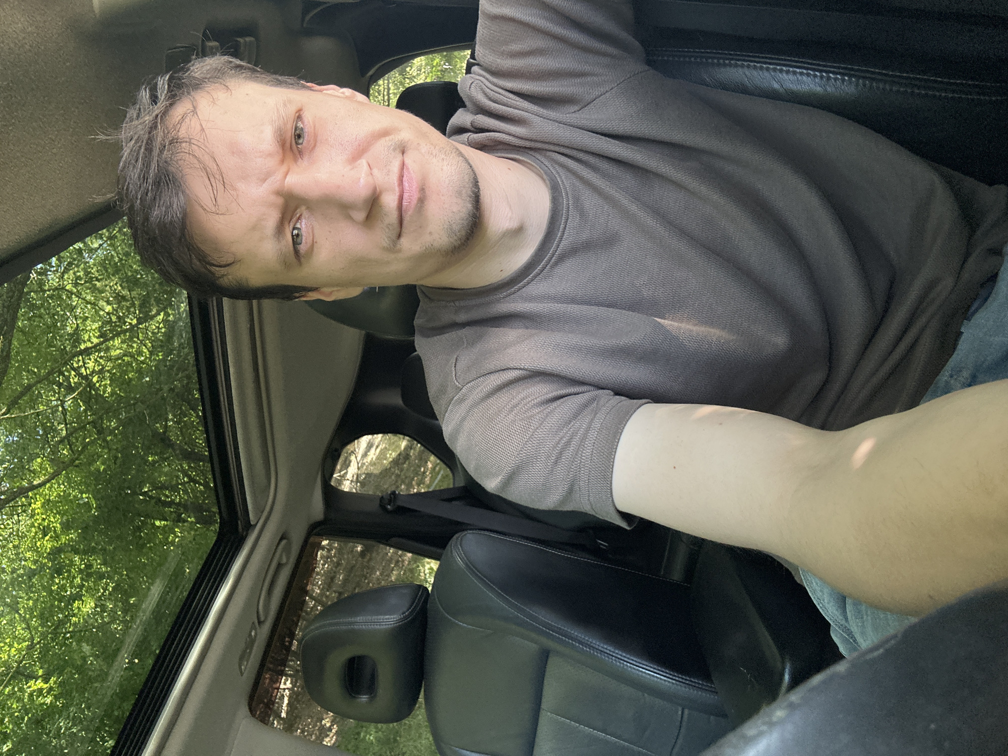 The image shows a man sitting in the driver's seat of a car, looking directly at the camera. The car has a sunroof open, revealing a canopy of green trees and foliage above. The man is wearing a short-sleeved, textured brown shirt and blue jeans. The interior of the car features black leather seats and a portion of the steering wheel is visible in the lower left corner. The man has short, dark brown hair and a light stubble on his face. The background outside the car is blurred, suggesting movement or a shallow depth of field, with the bright green of the trees indicating a sunny day. The image is a selfie, taken from the perspective of the driver. The location is Makiv, Ukraine, indicated by context outside the image.