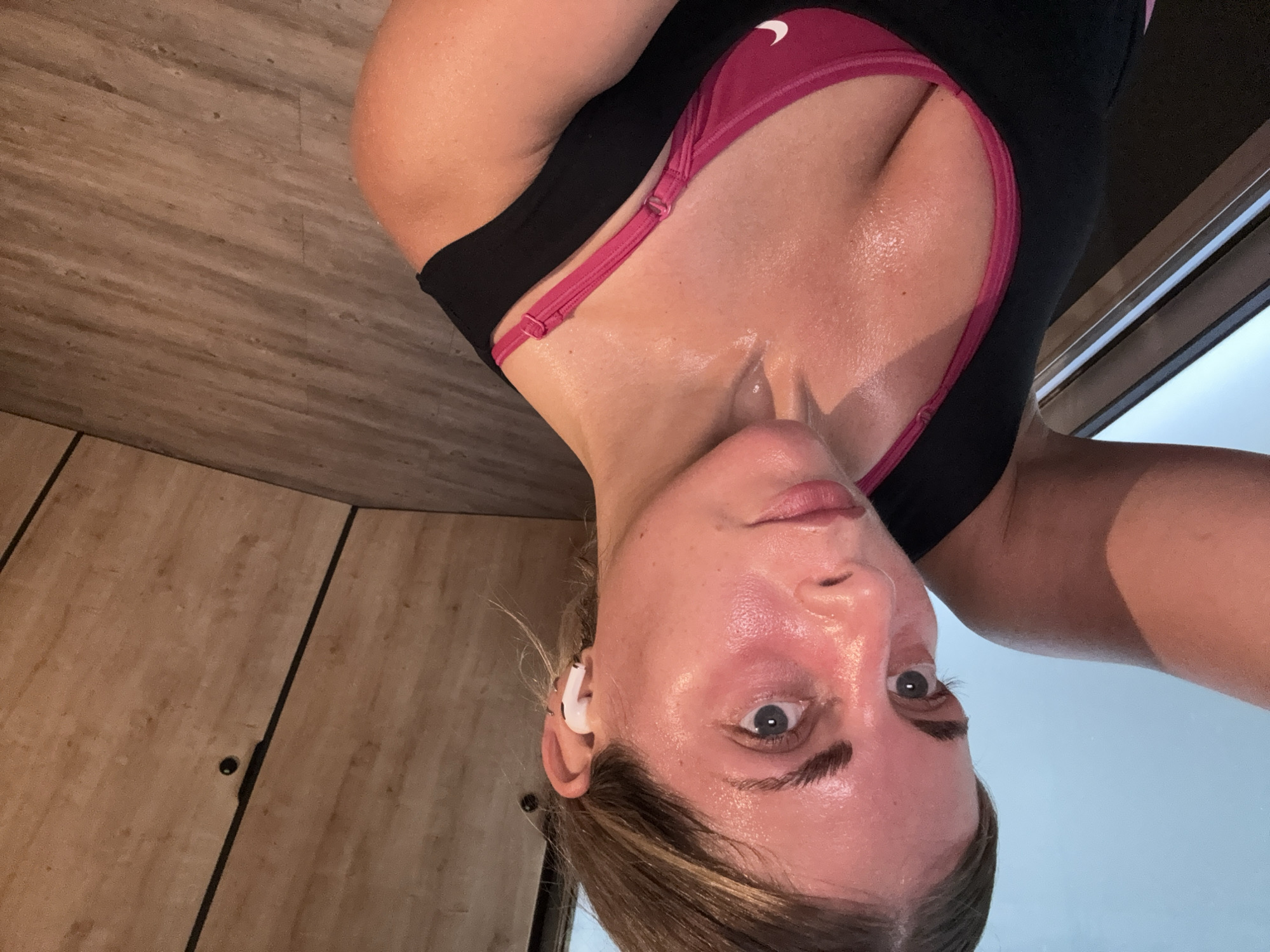 A selfie of a woman in workout clothes. The woman is wearing a black and pink Nike athletic top and white wireless earbuds. Her face is sweaty. The background includes a wood-paneled wall and a window. The image is rotated, making the woman appear upside down. The location is not Paris, France.