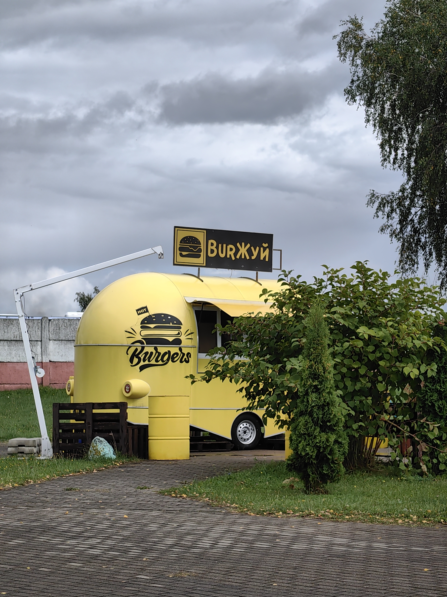 An overcast, cloudy day in Myadzyel, Belarus, features a bright yellow food truck shaped like a retro camper van. The truck has a large sign above it that reads "BurЖуй" in yellow Cyrillic letters on a black background, which translates to "Burger." The side of the truck displays a black graphic of a burger with the word "Burgers" in stylized script below it. Next to the truck, there is a small, green coniferous tree and a lush green bush. In front of the truck, a paved walkway with interlocking grey bricks leads towards it. To the left of the truck, a concrete fence is visible, and a wooden pallet structure is near the entrance. A metal arm extends upwards from the side of the truck, possibly for a light or sign. The ground is covered with grass and fallen leaves. The overall scene depicts a food vendor setup in an outdoor location.