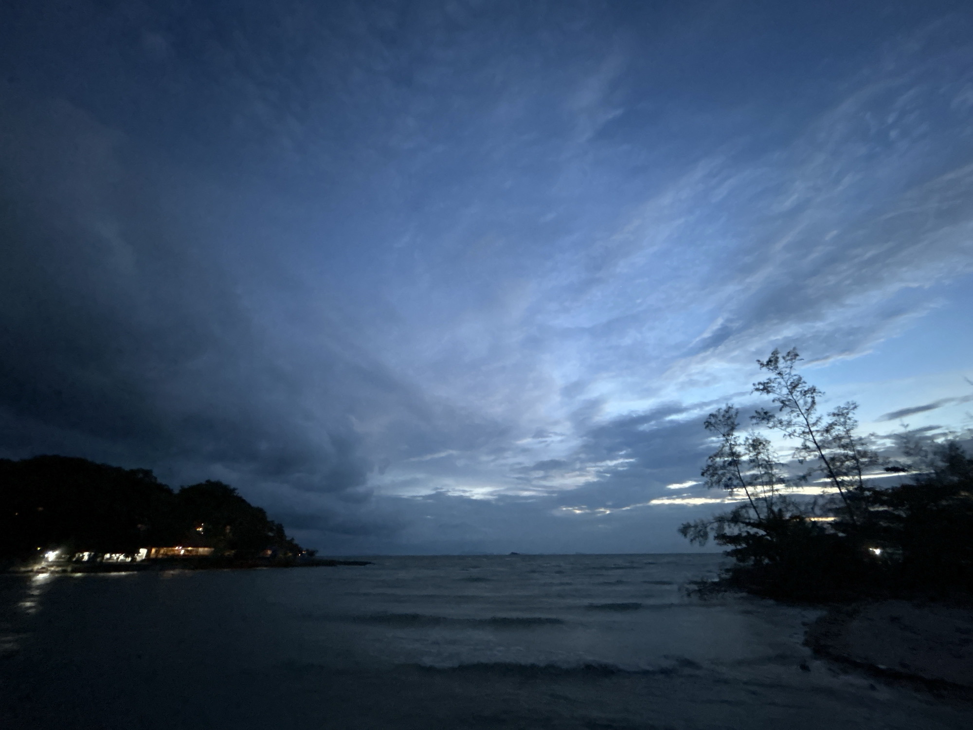 A wide shot captures the twilight over a beach in Ko Pha-ngan District, Thailand. The sky is a gradient of deep blue to lighter hues, with streaks of clouds moving across. The sea is a dark, choppy expanse, with small waves reaching the shore.
On the left side of the frame, a dark, tree-covered hill is visible, dotted with the warm glow of lights from buildings nestled amongst the trees, suggesting a village or resort. The lights reflect faintly on the dark water near the shore.
On the right side, silhouetted trees and foliage stand against the lighter sky, with a patch of sand in the foreground. The overall atmosphere is tranquil, with the end of daylight transitioning into night. No people are visible in the frame.
