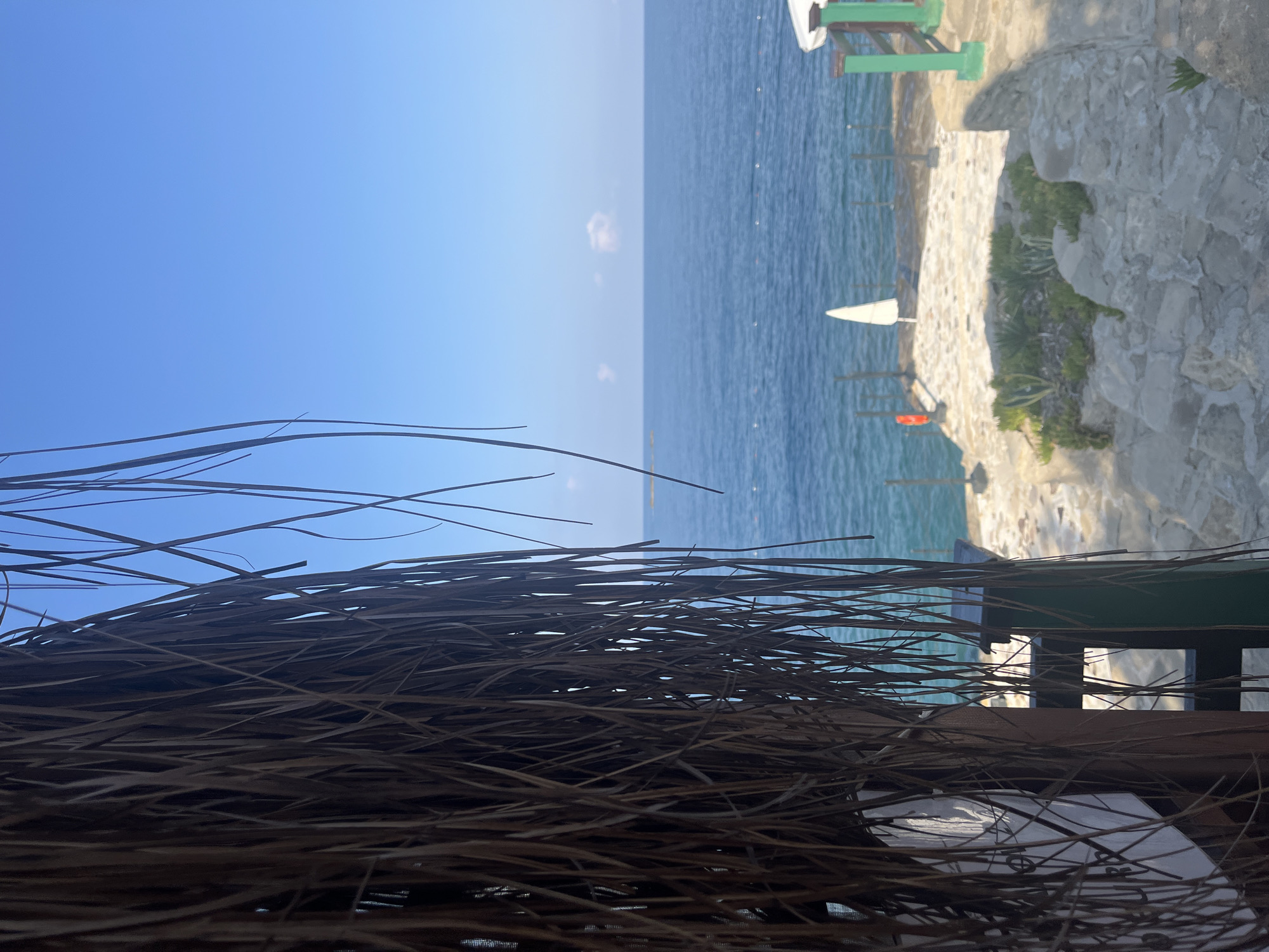 A view from under a thatched roof looks out onto the Mediterranean Sea under a clear blue sky. The foreground is dominated by the dried grass of the roof, with thin strands hanging down. Beyond the roof, a stone pathway leads to a wooden railing overlooking the water. A white umbrella is visible on the pathway. The sea is a deep blue, with a few small white buoys marking a boundary in the distance. A small, rocky island is visible on the horizon. The scene appears to be a resort or beach area in Alanya, Turkey.
