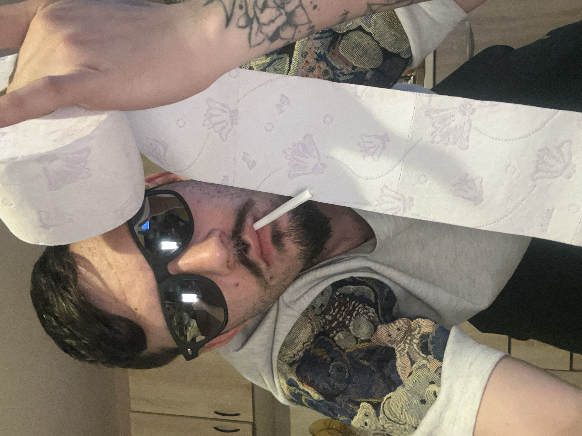An image, rotated 90 degrees clockwise, presents a close-up of a man from the chest up, in what appears to be an indoor setting in Varna, Bulgaria. He has dark hair, a mustache, and a short beard, and is wearing dark sunglasses that reflect light. A cigarette is positioned in his mouth.
The man is dressed in a light grey hooded sweatshirt, with the sleeve on his left arm featuring a detailed, tapestry-like pattern in shades of dark blue, green, and brown. Tattoos are visible on his left arm, extending from beneath the sleeve.
A roll of white toilet paper is prominently displayed, unrolling diagonally across the upper portion of the frame, partially obscuring the man's head and the background. The toilet paper has a distinct light purple/lavender pattern consisting of stylized floral or butterfly motifs interspersed with small dotted circles and lines. The background behind the man is blurred, suggesting an indoor environment with light-colored elements. No visible text is present in the image.