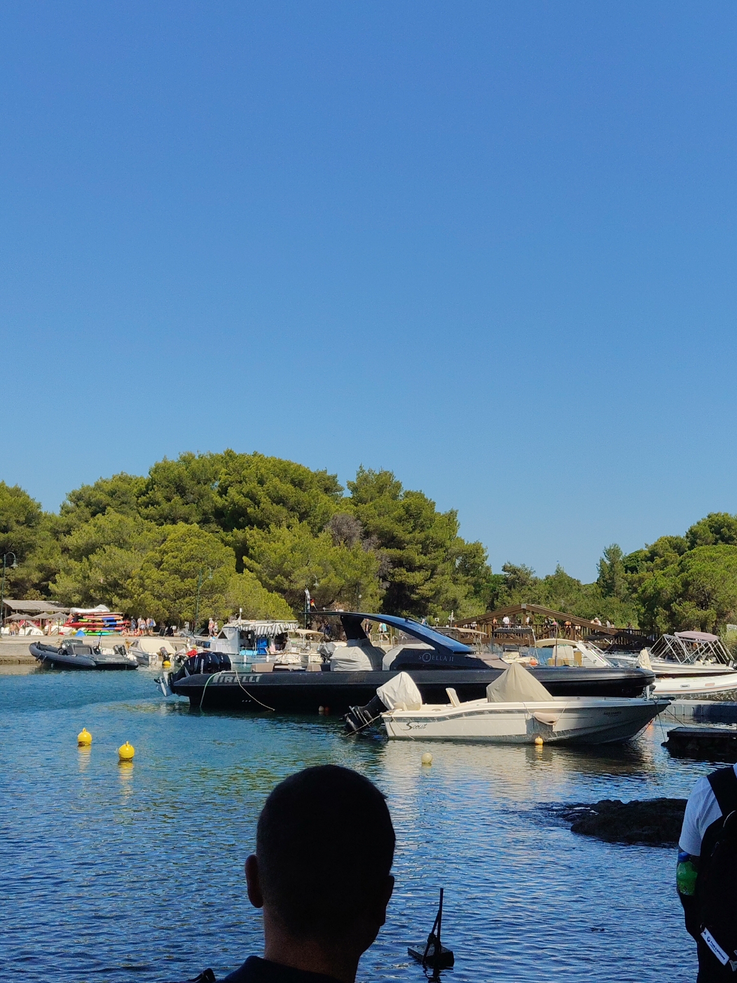 The image depicts a sunny day at a harbor in Greece, with clear blue skies and calm waters. Several boats are docked, including a sleek black motor yacht named "STELLA II" and a smaller white boat with "Scout" written on its side. In the background, a lush green forest covers a hillside, with a few structures and umbrellas suggesting a beach or recreational area. People are visible on the shore and on some of the boats, indicating a lively atmosphere. In the foreground, the back of a person's head is visible, looking out at the water. The scene is peaceful and picturesque, capturing a typical summer day in a Greek coastal town.
