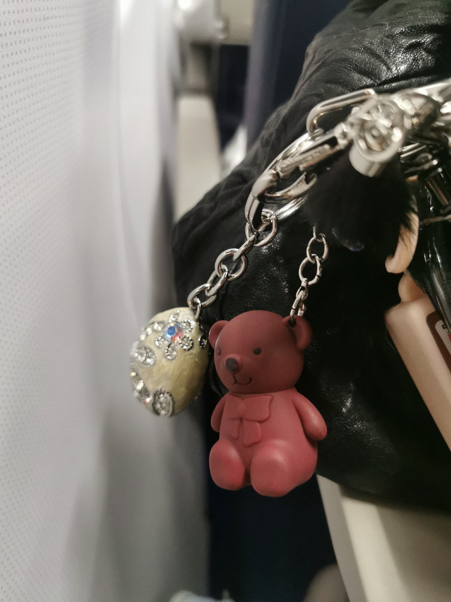 The image shows a close-up view of a black leather bag with several keychains attached. One keychain features a plush, red-brown teddy bear with a scarf. Another keychain is an oval-shaped charm adorned with rhinestones and a central blue gem. The chains and clips connecting the keychains are silver-colored. The background is blurred, showing a white, textured surface and a hint of blue, suggesting an interior space, possibly an airplane cabin. The location, Новое Мартьяново, Russia, is not discernable from the image.