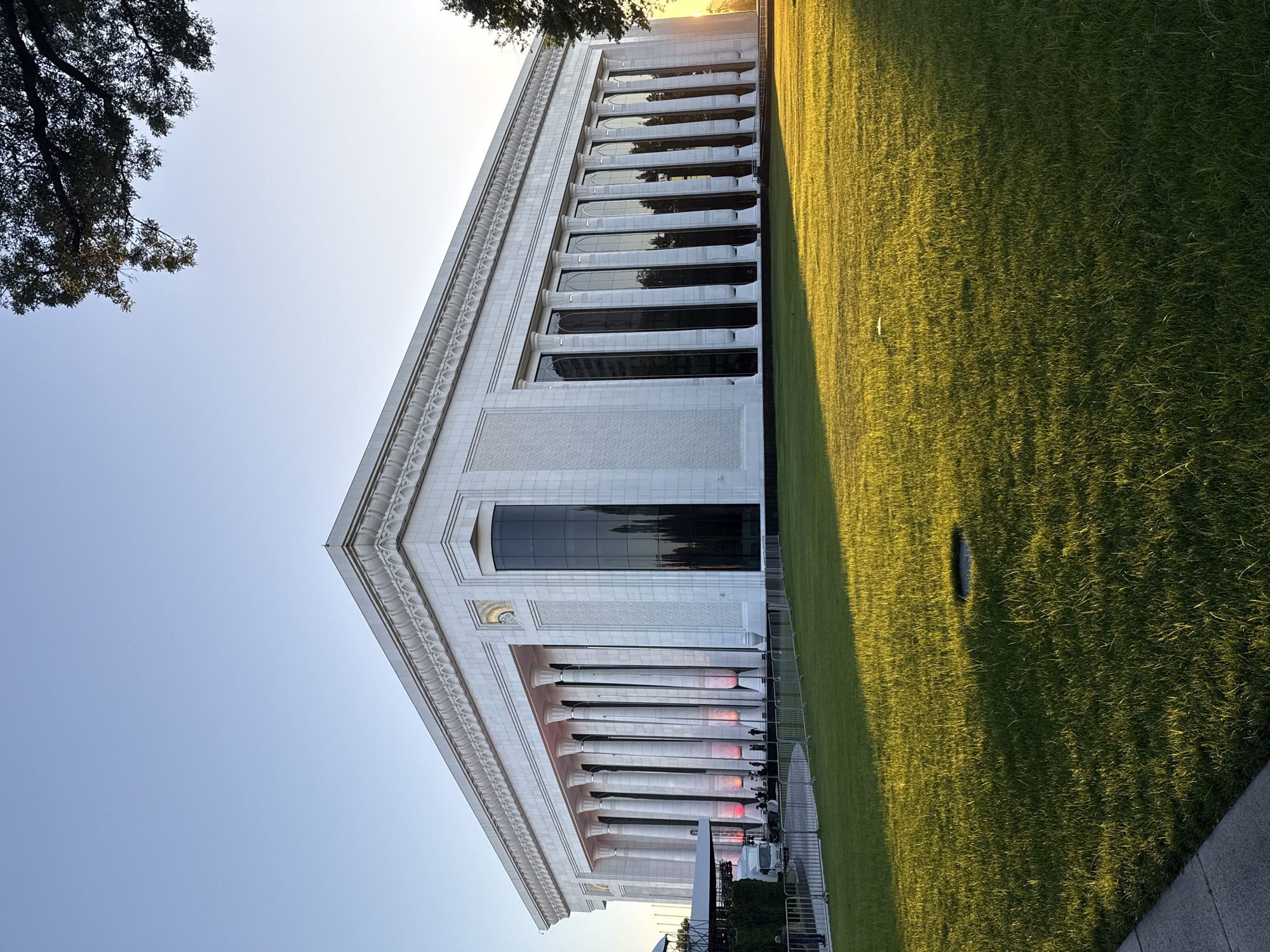 The image shows a building in Tashkent, Uzbekistan, likely a government or administrative building, judging from its architectural style with classical columns and a triangular pediment. The building is constructed of light-colored stone or concrete. It has several floors with long, vertical windows. A large lawn stretches along the side of the building, casting a shadow on the green grass. The sky is a clear, light blue, suggesting either early morning or late afternoon. Trees are partially visible on the periphery of the image. Construction or filming equipment and barriers are set up near the base of the building, suggesting an event or activity is taking place. The city is not discernible from this vantage point.
