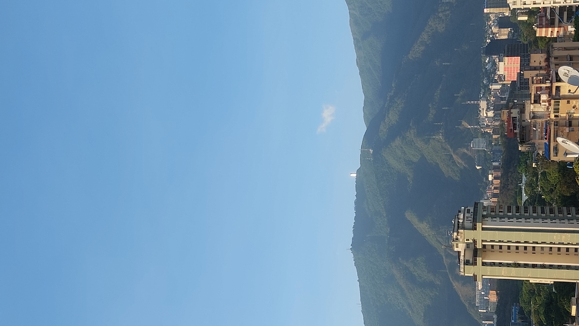 The image shows a view of the city of Caracas, Venezuela, featuring a clear blue sky taking up approximately half of the frame. A mountain range with lush green vegetation runs vertically down the right side of the image. At the base of the mountain, there is a dense urban area with residential and commercial buildings visible. A tall, light-colored building with multiple floors stands out in the lower right corner of the frame.