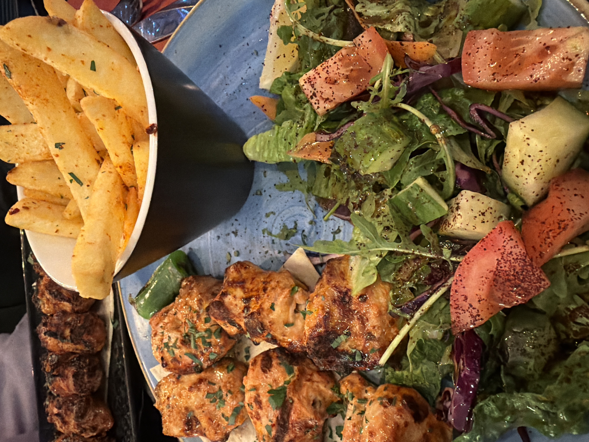 The image captures a plate of food consisting of french fries in a black conical container, a salad with tomatoes and cucumbers, and grilled chicken skewers. The food is likely in a restaurant setting. While the image provides no specific clues to confirm the location as London, United Kingdom, it is plausible given the widespread availability of similar cuisines in the city. The environment appears to be dimly lit, suggesting an indoor setting, possibly in the evening. There are no people visible in the image.