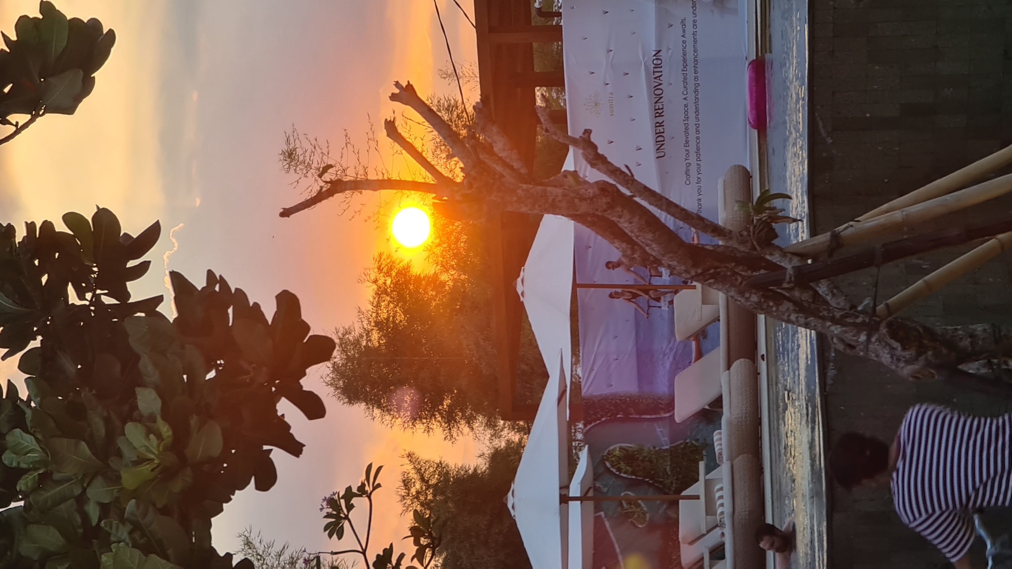 A resort swimming pool is seen at sunset in Denpasar, Indonesia. The sky is filled with warm orange and pink hues from the setting sun, which is a bright orb in the middle of the frame, partially obscured by trees. In the foreground, lush green leaves hang from a tree branch. A bare, twisted tree trunk with bamboo poles supporting it stands in front of the pool.
The pool itself is a clear blue, with lounge chairs and white umbrellas lining the edge. In the background, a large banner reads "UNDER RENOVATION" and "sumitra", with a message about crafting an elevated space. Two people, a man and a woman, are visible on stationary bikes beside the pool. A man is swimming in the pool, and another person is visible in the foreground with their back to the camera, wearing a striped shirt. A bright pink inflatable ring is floating in the water.