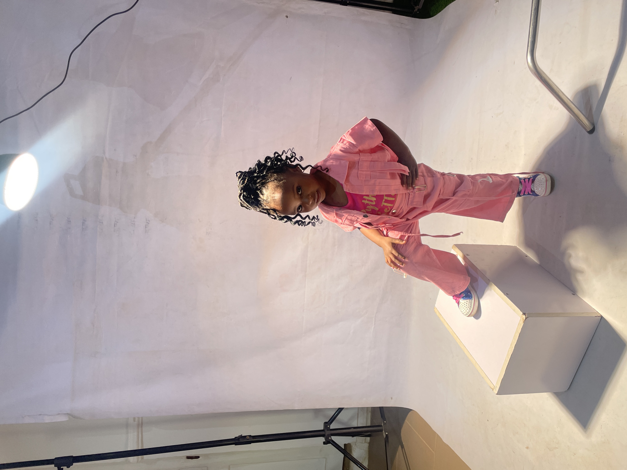 A young girl with dark hair styled in braids is standing on a white box in what appears to be a photo studio. She is wearing a pink outfit with a matching pink t-shirt underneath, and colorful sneakers. She is looking directly at the camera with a slight smile. The background is a white backdrop that is draped and shows wrinkles. A lighting fixture is visible on the left side of the frame, casting bright light on the scene. Portions of what seems to be studio equipment are also seen in the lower area of the frame. The location is likely in the city of Barakin Akawo, Nigeria.
