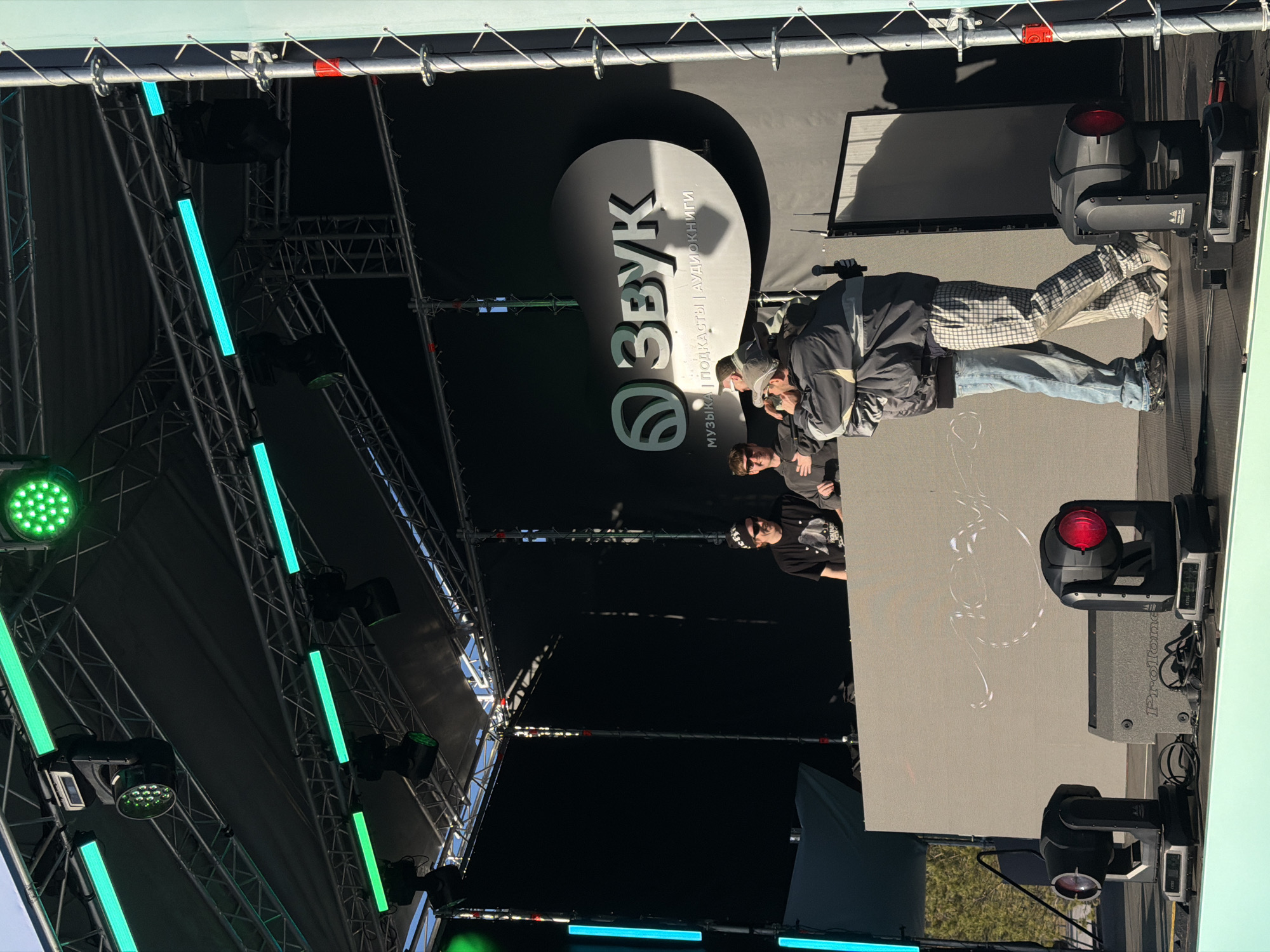 A stage is set up, possibly outdoors, under a grid-like structure supporting lighting equipment. A sign for "Звук" (Sound) is visible in the background. People are present on the stage. The location is possibly Ozyornoye, Russia, based on the Cyrillic signage. There is a grey screen or wall on the right side of the stage with abstract white markings. Red and green lights are visible in the rigging above the stage.