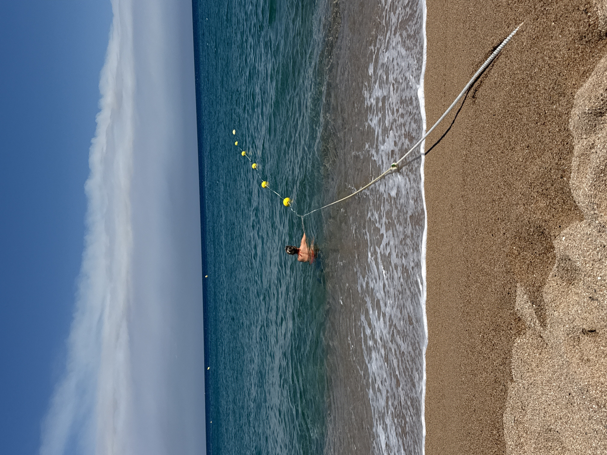 The image shows a view of the Mediterranean Sea from the beach in Blanes, Spain. The foreground is a sandy beach with gentle waves lapping the shore. A thick rope, possibly a mooring line, extends from the water onto the sand at an angle. Further out in the turquoise water, a person wearing a red bathing suit is swimming. A line of yellow buoys stretches across the sea in the mid-ground, marking a boundary. The sky above is a clear blue with scattered white clouds. The overall scene depicts a tranquil day at the beach.