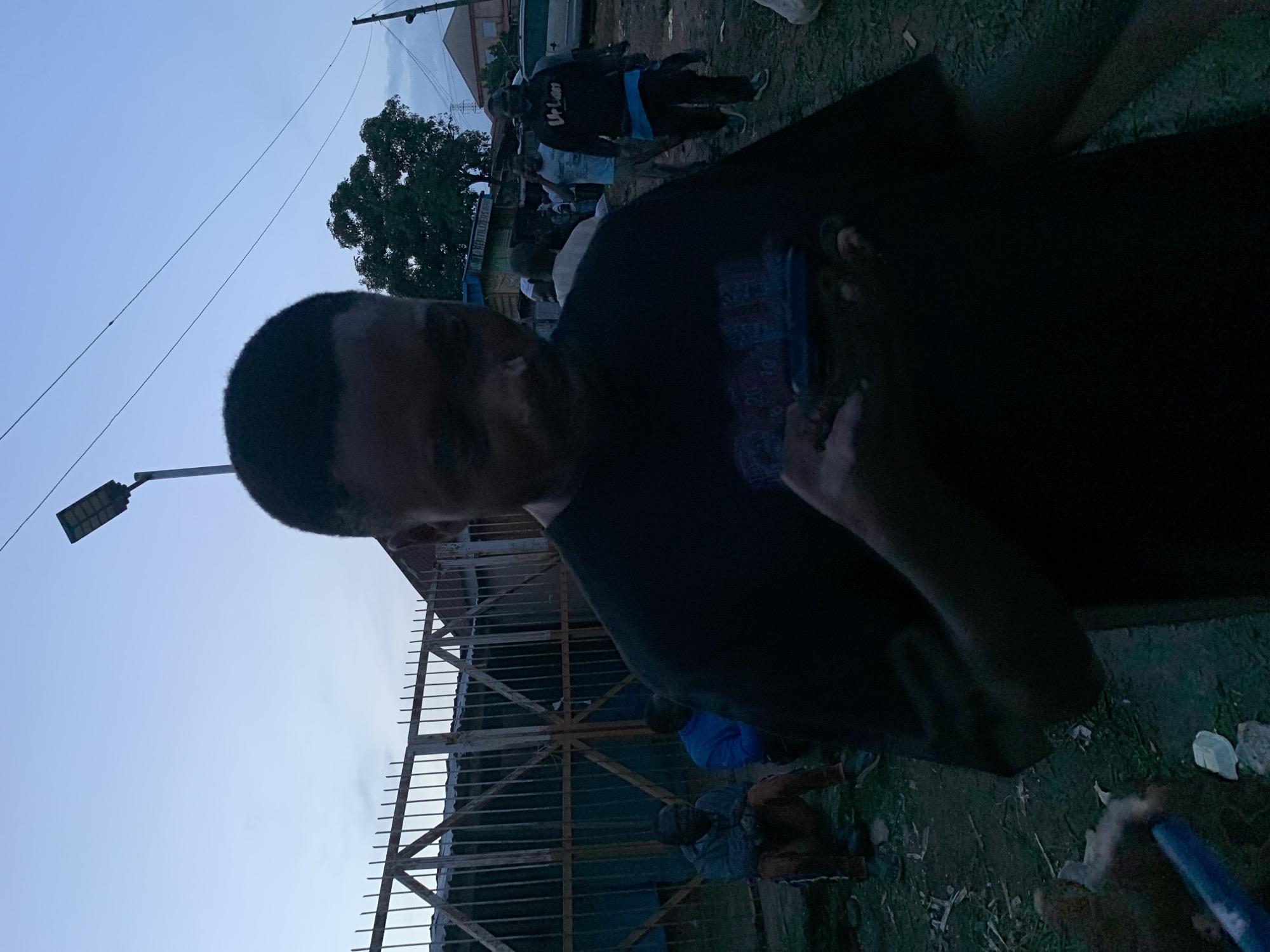 The image depicts a street scene in Gura Top, Nigeria, during twilight. In the foreground, a young man is looking down at his phone. He is wearing a black t-shirt with a graphic design on it. To his left, two men are seated on the ground near a metal fence. The man on the left is wearing a grey jacket and orange pants, while the man on the right is wearing a blue top. In the background, other people are visible walking and standing, and there are several buildings and a street light. The ground is covered with dirt and some vegetation.