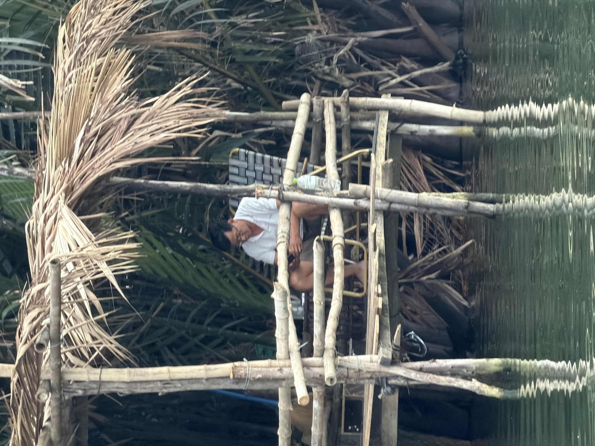 An upright, eye-level shot captures a man sitting on a rudimentary wooden and bamboo structure built over a body of water, likely a river or canal, in Hội An Ward, Vietnam.
The man, wearing a white short-sleeved shirt and dark shorts, is positioned centrally, leaning forward with his hands on the bamboo planks. He appears to be resting or observing the water below. A clear plastic water bottle with a light-colored cap rests on the structure next to him. He is using a simple chair or stool with a black and white checkered fabric seat.
The structure itself is made from rough, unpolished bamboo poles and logs, lashed together to form a platform or small pier. Dense, tropical vegetation surrounds the scene. To the man's left and behind him are prominent, dried, light-brown palm fronds, while darker green foliage creates a lush backdrop.
The water, visible in the lower part of the frame, is dark green and exhibits distinct, irregular ripples and reflections, suggesting gentle movement or light refraction. The lighting appears soft, indicating an overcast day, deep shade, or early/late daylight. No discernible text is visible on the bottle or any other object. The overall atmosphere suggests a quiet, natural, waterside setting.