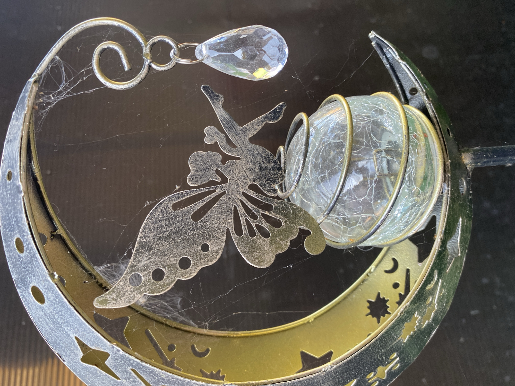 The image shows a close-up of a decorative solar garden stake featuring a crescent moon with celestial cutouts, a fairy silhouette, a glass orb wrapped in wire, and a clear teardrop-shaped crystal. The stake is designed to be placed in the ground outdoors. The metal elements are silver and gold-toned, with some signs of weathering and the presence of cobwebs. The background is a dark, blurry surface, likely a wall or fence, suggesting an outdoor setting. The scene is static, with no people or actions depicted. The composition focuses on the details of the ornamental object, highlighting its craftsmanship and textures. The location of this object in Nizhniy Lomov, Russia is not discernible from the image itself.