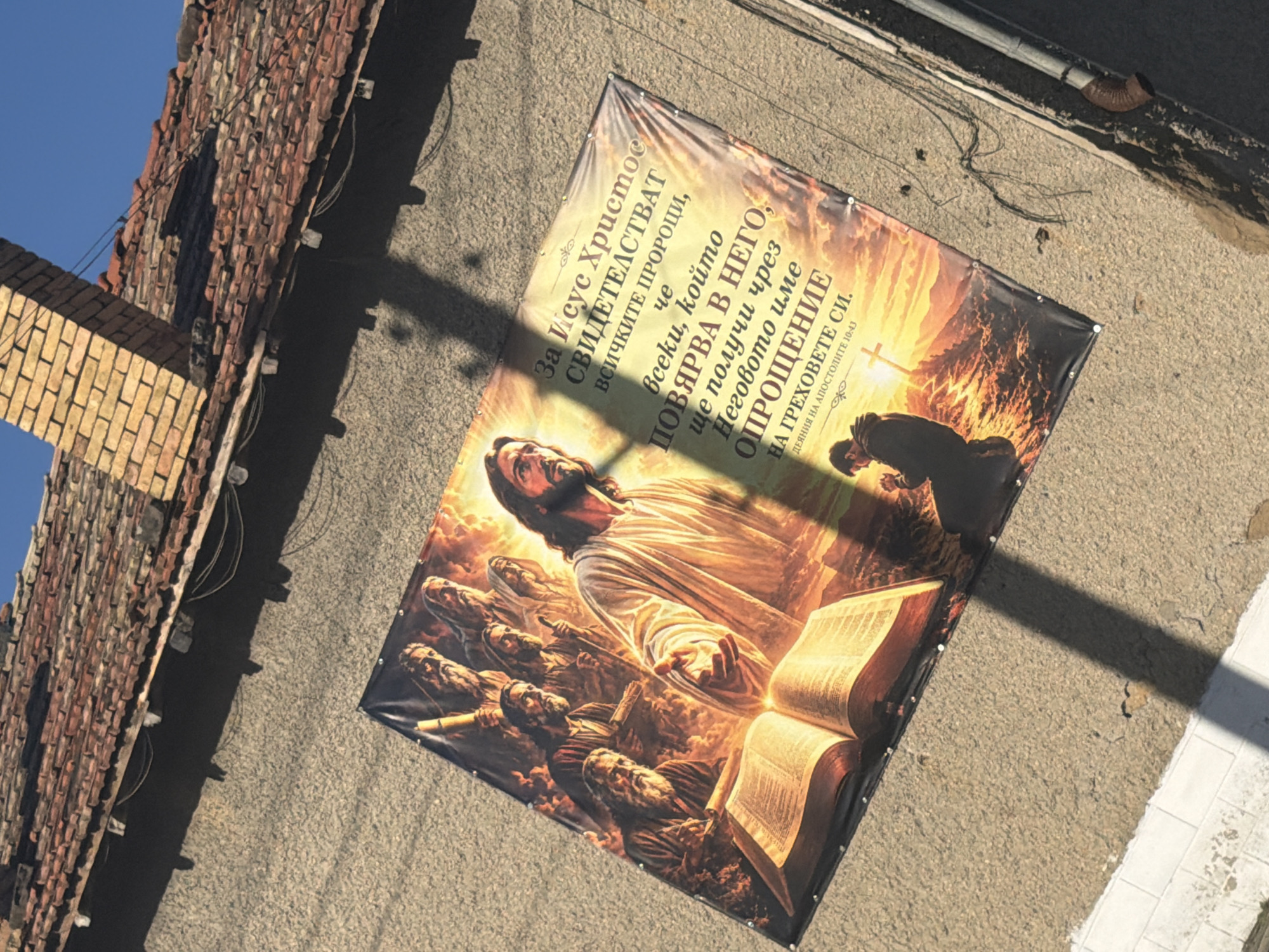 A large banner displaying a religious scene hangs on the side of a building with a red-tiled roof. The banner depicts Jesus Christ in the center, surrounded by other figures. An open book rests in the foreground. To the right of the figures, text in Cyrillic script is visible. Below the main scene, a person is shown kneeling in prayer with a cross in the background. The building appears to be made of concrete with visible texture. The roof has some missing or discolored tiles and is partially shaded by a brick chimney. Electrical wires are visible attached to the roofline and running along the side of the building. The image was taken in the city of Veselinovo, Bulgaria, as indicated by the text on the banner: "За Исус Христос свидетелстват всичките пророци, че всеки, който повярва в него, ще получи чрез неговото име опрощение на греховете си. Деяния на Апостолите 10:43". This translates to: "All the prophets testify of Jesus Christ that everyone who believes in him will receive through his name forgiveness of sins. Acts 10:43".