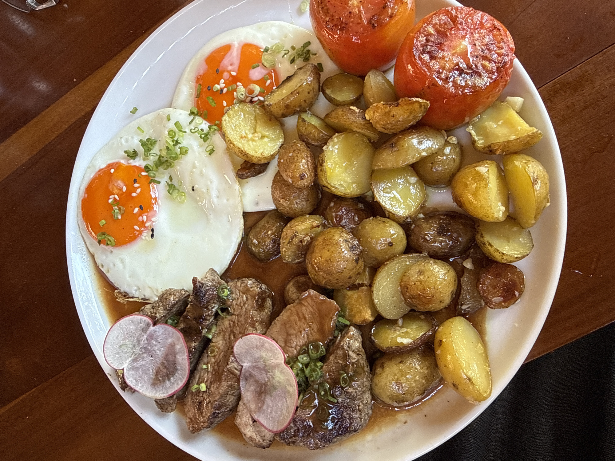 A top-down, close-up shot depicts a white plate filled with food. The dish consists of two fried eggs topped with green onions and white sesame seeds, roasted potatoes, two roasted tomatoes, and sliced meat garnished with radish slices and green onions, all sitting in a sauce. The plate is placed on a wooden surface. The environment and food style suggest the location could be in the city of Kuta Selatan, Indonesia.