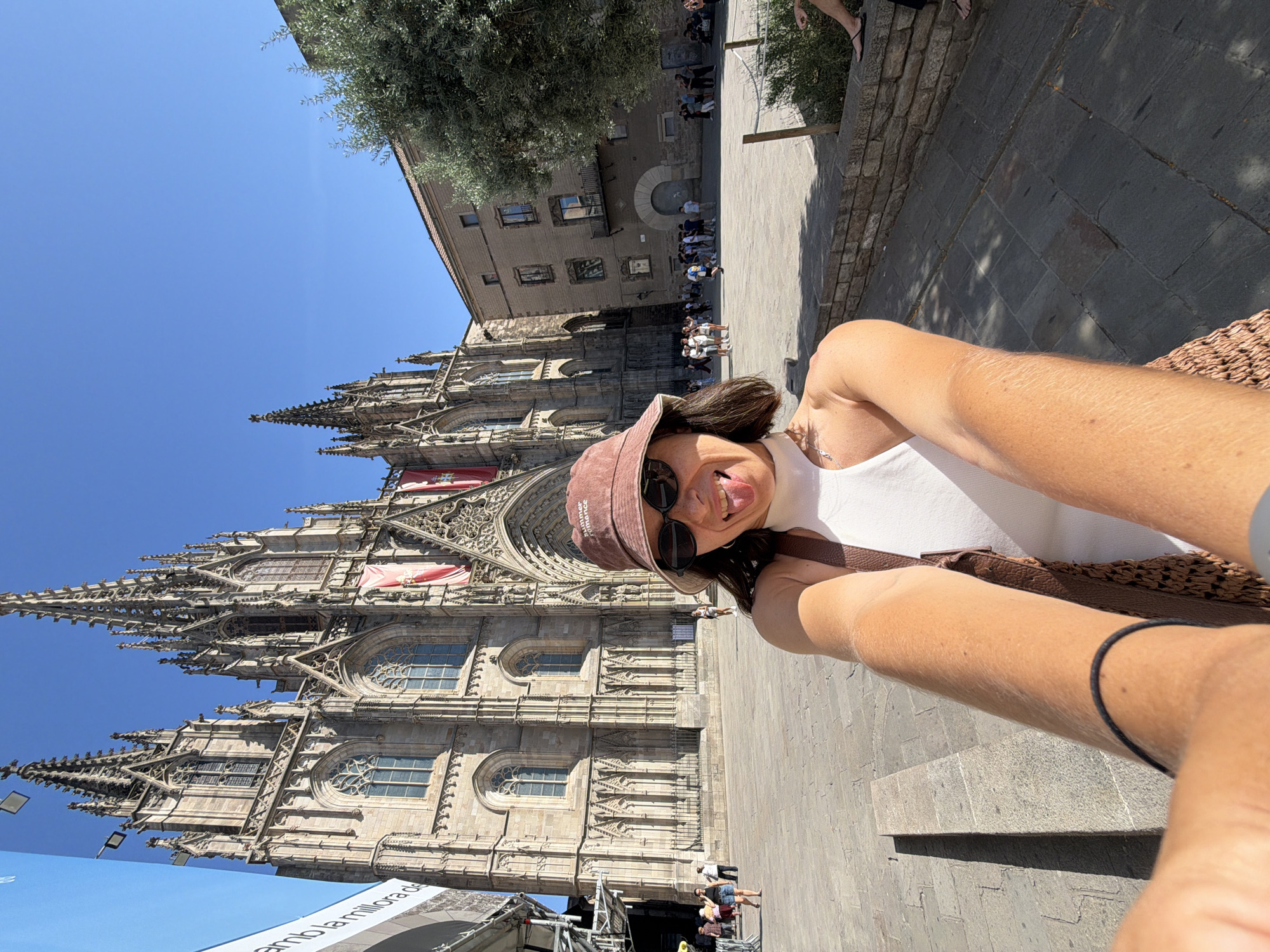 A woman with dark hair, wearing a bucket hat and sunglasses, is taking a selfie in front of the Barcelona Cathedral. She is sticking her tongue out and has a playful expression. She is wearing a white tank top and a woven bag. In the background, the impressive facade of the cathedral, with its Gothic architecture and spires, is visible under a clear blue sky. Tourists are scattered around the plaza in front of the cathedral, some walking and others observing the architecture. An olive tree is to the right of the frame.