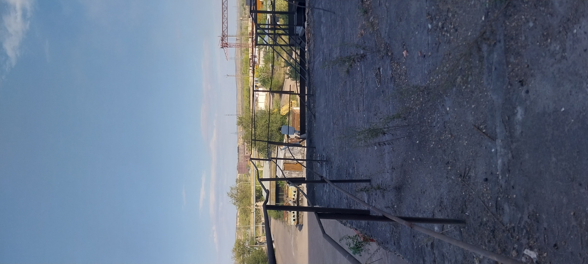 An outdoor, eye-level, vertical shot shows a dilapidated industrial landscape under a clear blue sky with some faint clouds on the horizon. In the foreground, a dark, textured surface, possibly a rooftop or ground, is littered with small pebbles and patches of dry vegetation. A rusty metal railing, with vertical posts, runs along the left side of the frame, extending into the distance.
Beyond the railing, an industrial area is visible. There are various structures, including what appear to be small buildings with weathered walls and doors, as well as metal frames and what might be discarded industrial equipment. In the mid-ground, there are trees and more industrial buildings in the distance. A prominent red crane is visible on the right side of the mid-ground, and power lines and pylons can be seen in the far distance, suggesting an industrial or power generation facility. The overall impression is one of disuse and decay within an industrial setting.