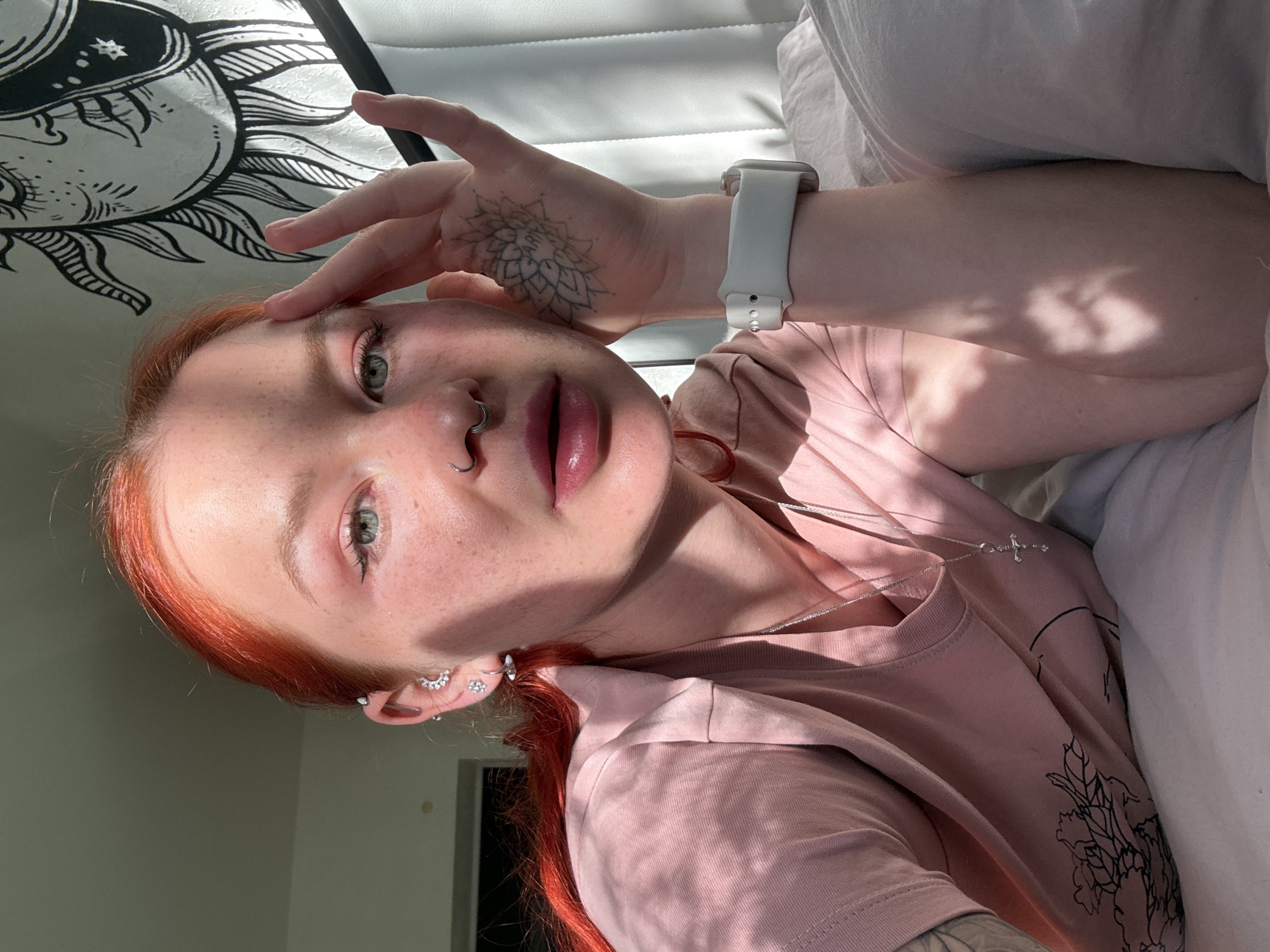 A woman with fair skin and freckles, red hair, and light green eyes is lying down. She has a silver nose ring and multiple earrings in her left ear. She is wearing a light pink t-shirt with a black floral design on it and a silver necklace with a cross pendant. On her left wrist, she is wearing a white smartwatch. She has a tattoo on her left hand. Her left hand is raised to her forehead. She is lying on a white pillow or cushioned surface. Behind her is a white tapestry with a black sun design. The location cannot be determined from the image.