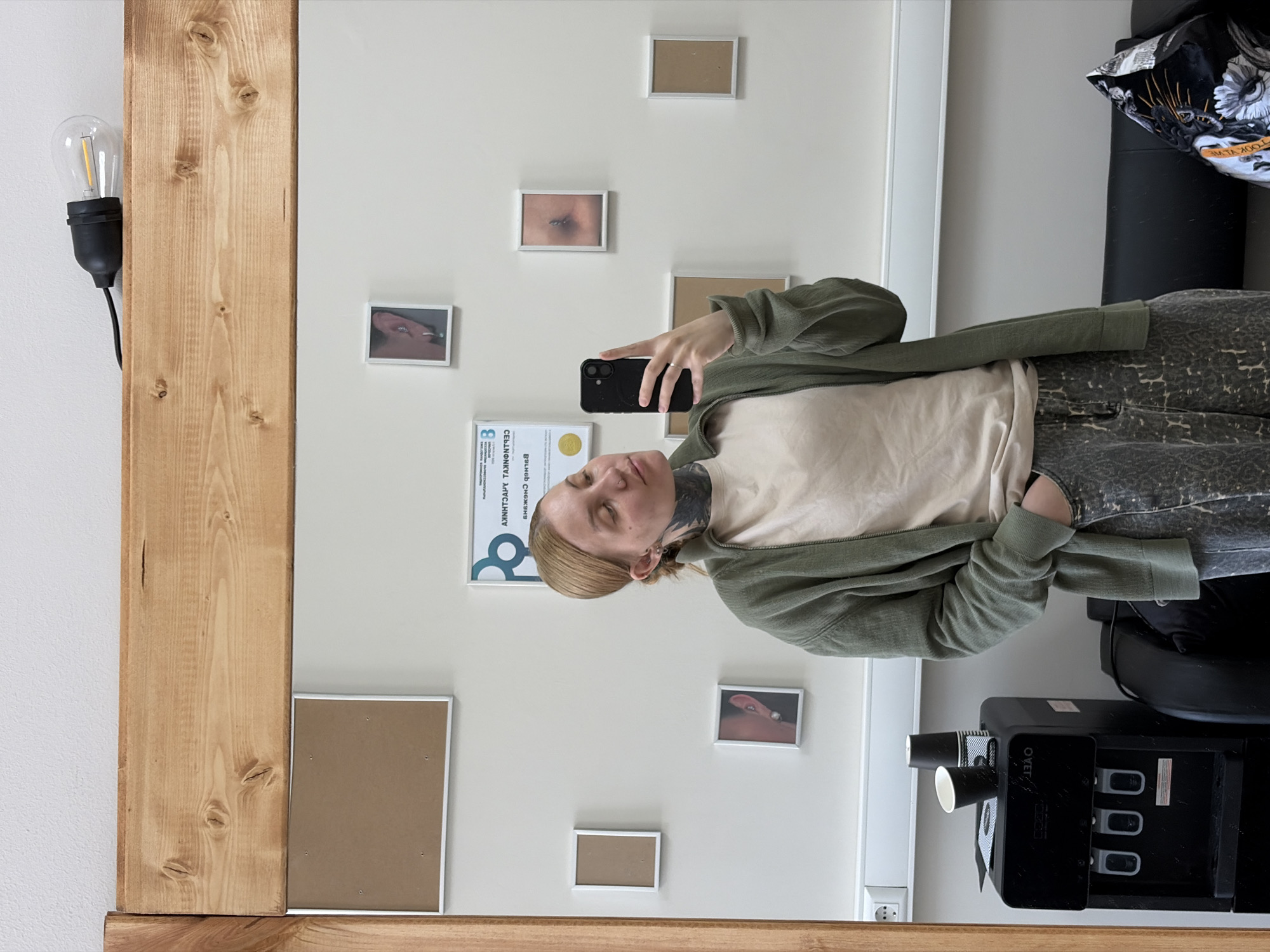 A person is taking a selfie in a mirror. The mirror has a wooden frame and a light bulb attached to its left side. The person is wearing a beige shirt and a green cardigan. Behind the person, on the wall, are several framed pictures and a certificate in Russian. A coffee machine is on the right side of the frame, along with a black leather chair. The location is Chaikovsky, Russia.