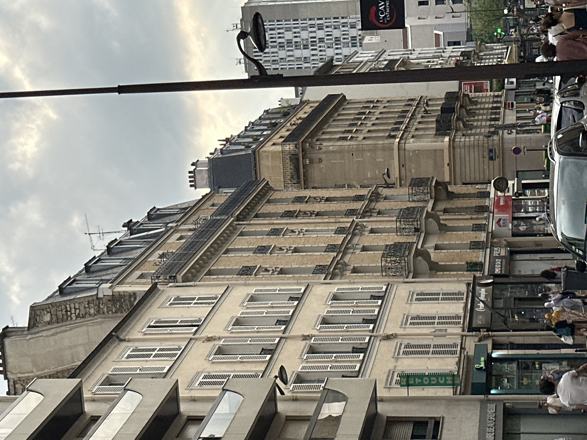 The image depicts a street scene in Paris, France, likely in a residential and commercial district. The architecture is characterized by multi-story buildings with classical facades, featuring mansard roofs, balconies with ornate railings, and numerous windows with white shutters. The buildings are closely packed, creating a canyon-like effect. In the foreground, a lamppost with an arm extending to the left is prominently featured. Several cars are parked along the street, and a few pedestrians are visible, some walking, others conversing. The shops on the ground floor of the buildings display signage in French, indicating businesses like "RETOUCHE" and other retail establishments. In the background, a taller, more modern building with a grid-like facade stands out against the overcast sky. The overall atmosphere suggests a typical urban environment on an ordinary day.