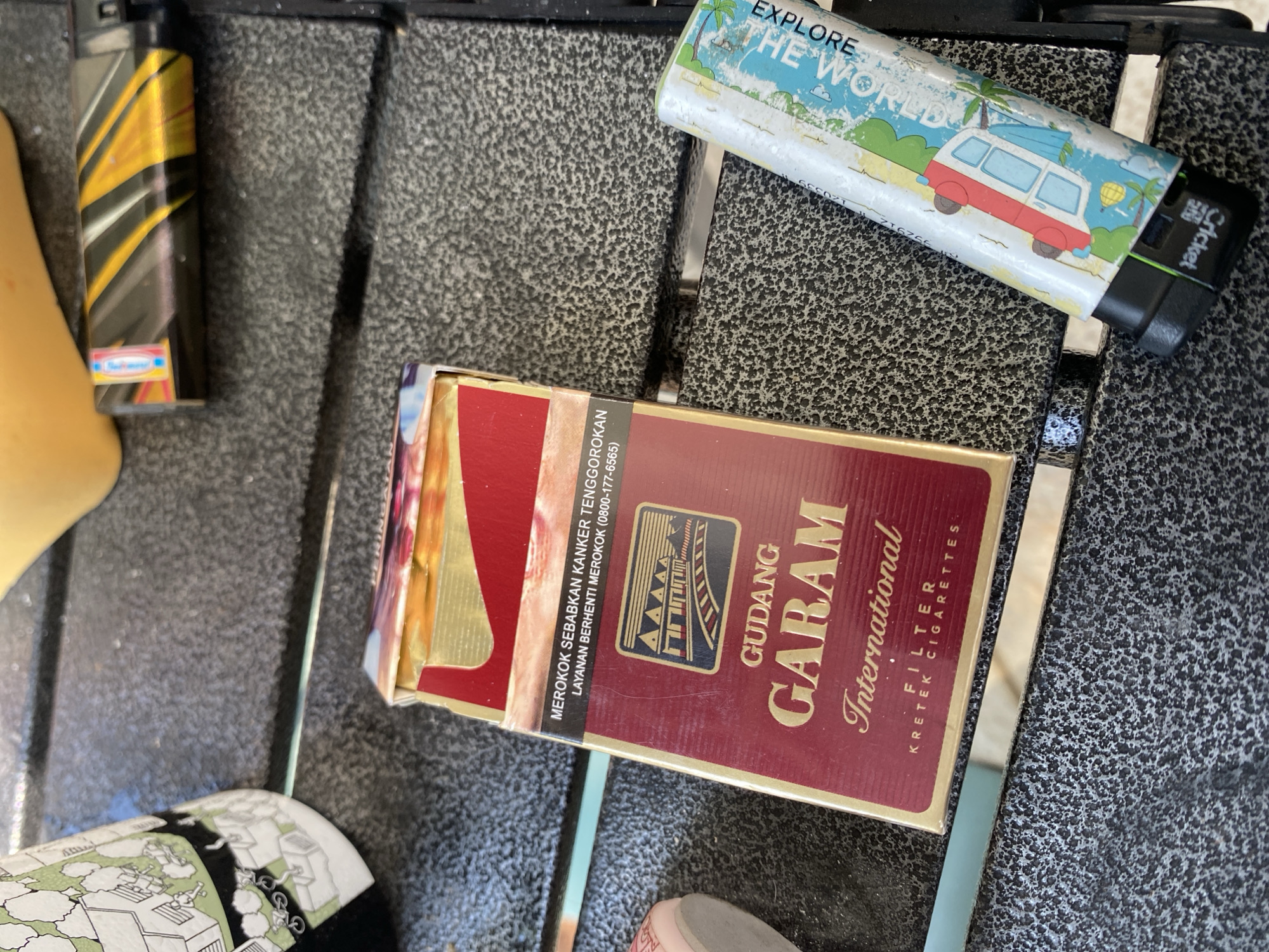 The image shows a close-up, overhead view of a rough, textured, dark gray surface, possibly a table or patio, with several objects resting on it. The main subject is a pack of Gudang Garam International filter kretek cigarettes, a popular Indonesian brand, positioned slightly off-center to the left. The pack is mostly maroon with gold lettering and a picture of a stylized train track and buildings. To the right of the cigarette pack is a lighter, its design featuring a colorful graphic of a camper van and palm trees with the words "EXPLORE THE WORLD" printed vertically. The lighter is identified as a "Cricket" brand by text on its base. In the upper left corner, a portion of a white object with a green and black illustrated design, possibly a can or container, is visible. In the upper right, a yellow, soft object, perhaps a cushion or piece of foam, is partially in frame. The lighting suggests it is daytime, with a bright, direct light source casting shadows. The scene does not contain any identifiable landmarks or specific clues that would indicate a precise location within Dukuh, Indonesia, beyond the presence of a well-known Indonesian product.