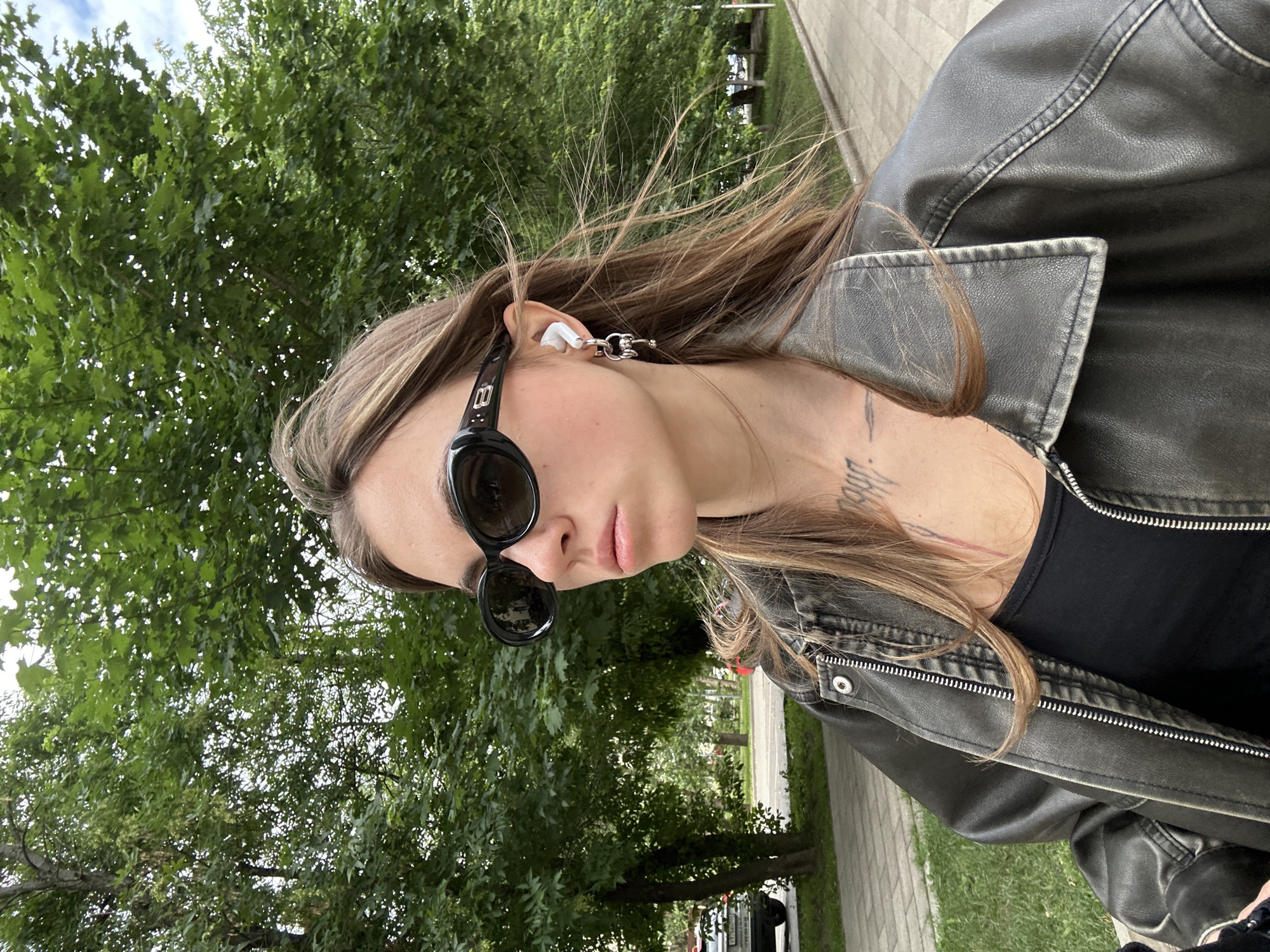 A young woman with long, brown hair wears black oval sunglasses and a black leather jacket over a black tank top. She has a white wireless earbud in her left ear and a silver earring with a charm in the same ear. She is looking to her left. Her skin is fair, and she has visible tattoos on her chest and neck. She is standing on a paved walkway next to a grassy area. In the background, there are numerous green trees, a street with cars, and a cloudy sky. The setting appears to be a park or a tree-lined street within the city of Moscow, Russia.