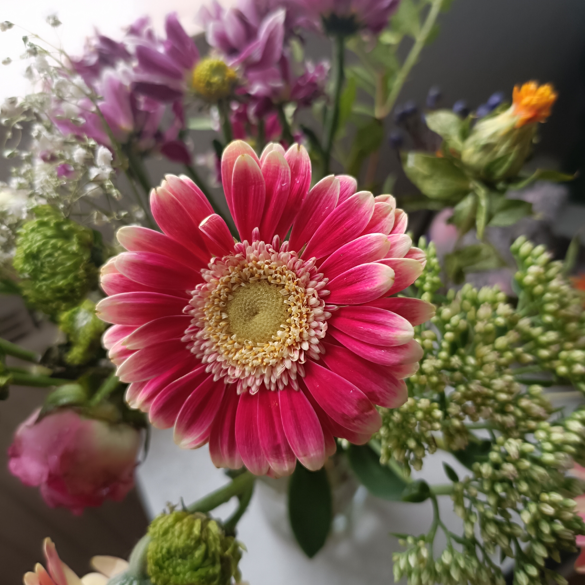 A close-up, high-angle shot focuses on a vibrant bouquet of flowers. The central subject is a large, bright pink gerbera daisy with a yellow center and delicate white-tipped petals. To the left and behind it are several out-of-focus pink chrysanthemums, a cluster of tiny white flowers, and a fuzzy green bloom. To the right, a bud with an orange tip and a branch covered in small, pale green buds are visible, along with some dark blue berry-like accents. The background is a soft, blurred mix of light and dark tones, suggesting an indoor setting. The image does not provide any information about Rüsselsheim am Main, Germany.