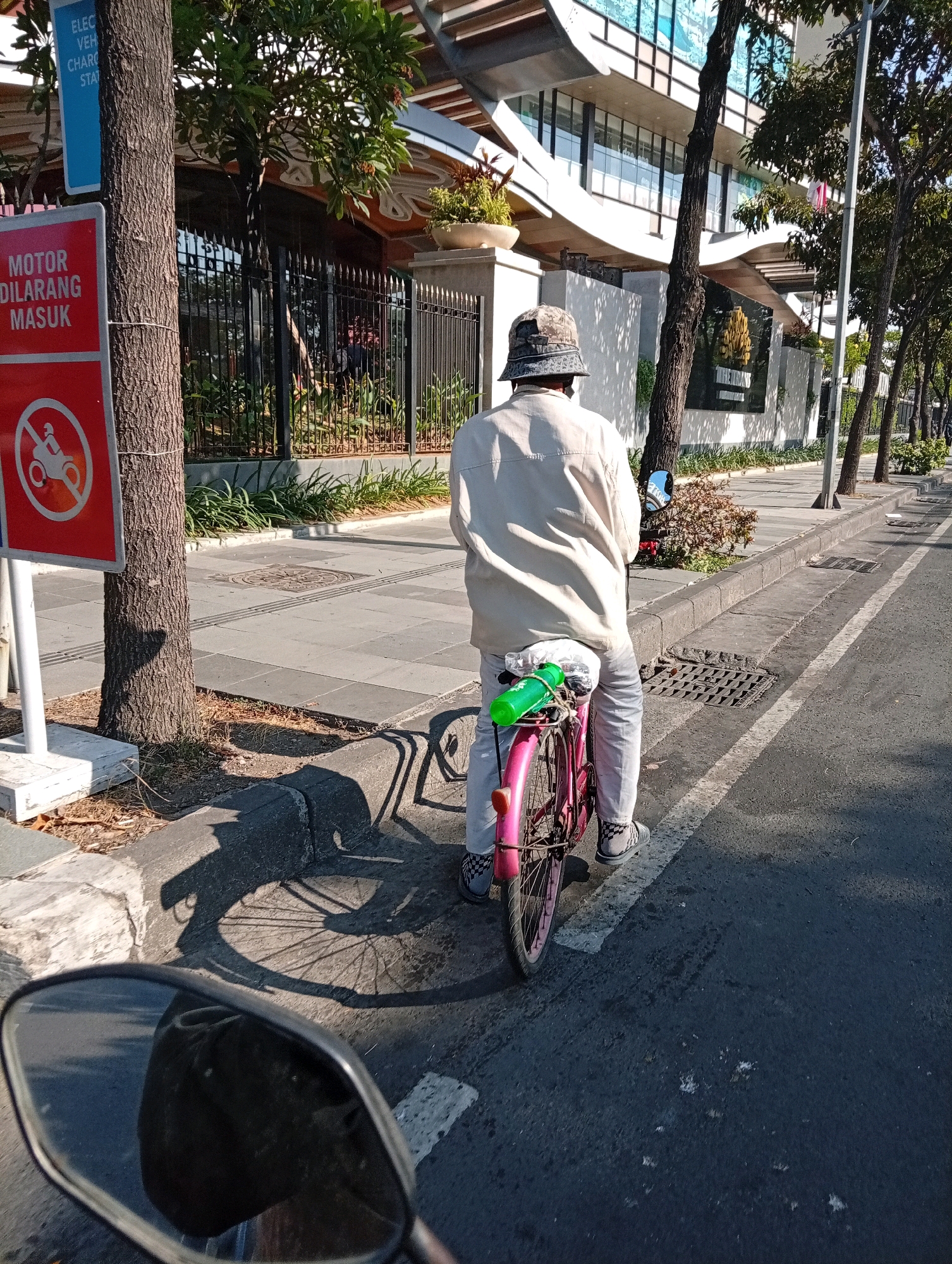 In Surabaya, Indonesia, a person is riding a pink bicycle on a paved road. They are wearing a light-colored jacket and light-colored pants, and a patterned bucket hat. The bicycle has a green water bottle attached to the back. The road is lined with trees and a sidewalk. A sign in Indonesian that reads "MOTOR DILARANG MASUK" meaning "MOTORCYCLES FORBIDDEN TO ENTER" is visible on the left side of the image. The background features a modern building with large windows. The scene is illuminated by sunlight, casting shadows on the road and sidewalk. The image appears to be taken from the perspective of a motorcycle due to the presence of a side mirror in the foreground.