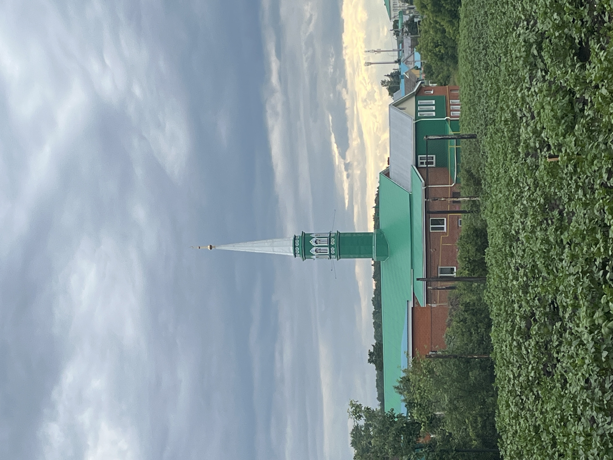 The image depicts a view from a field of crops, likely potatoes, towards a cluster of buildings. In the center, a prominent green minaret with a white spire topped with a golden crescent dominates the skyline. Adjacent to it, a brick building with a bright green roof and green trim is visible, featuring white-framed windows. To the right, another building with a gray roof and green trim, also with white-framed windows, stands alongside other colorful structures in the distance. The sky is overcast with heavy, dark clouds, but a sliver of lighter sky with yellowish hues can be seen towards the horizon. Trees are present on the left and right sides of the image, framing the buildings. The overall scene suggests a rural or semi-rural setting within the city of Bolshoy Kukmor, Russia. There are no people or animals visible in the frame.