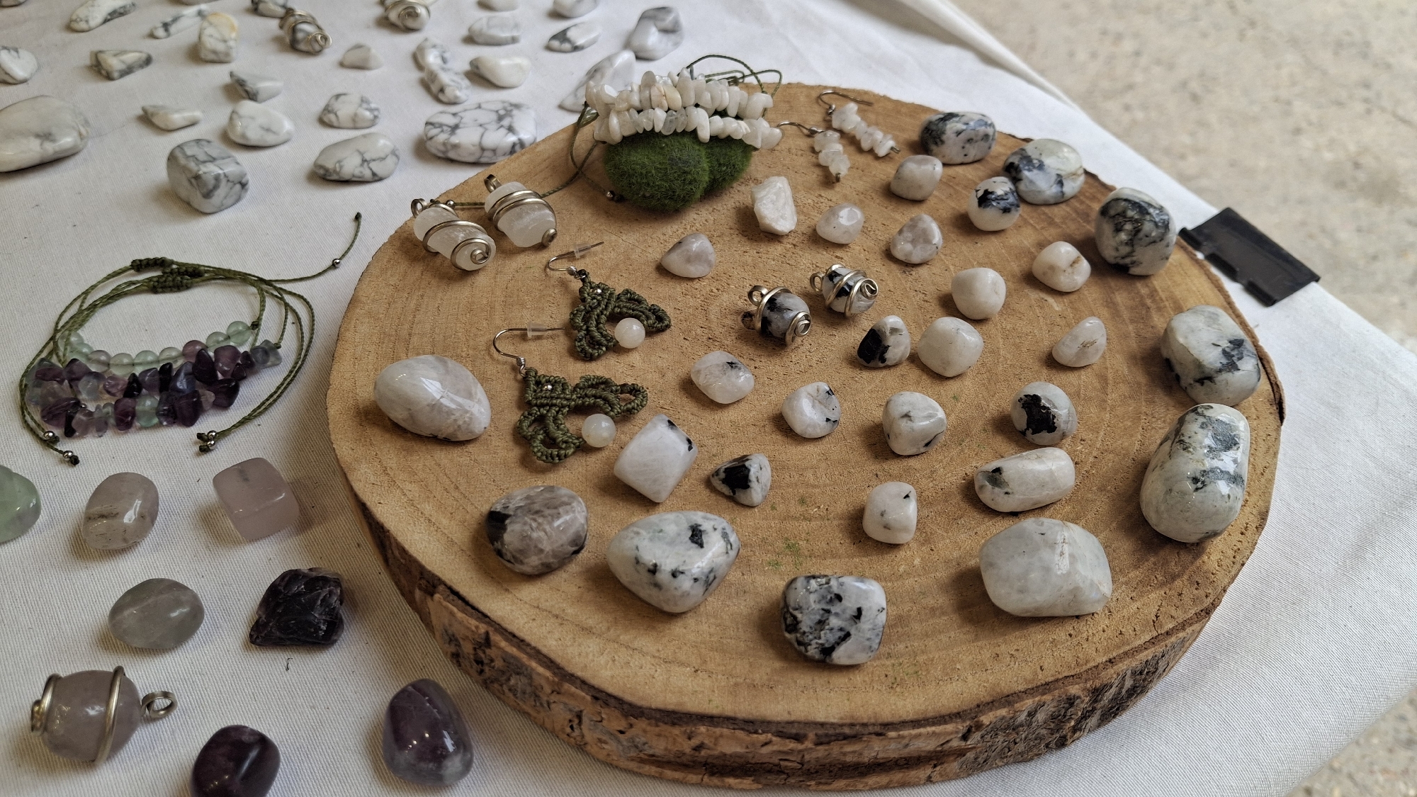 The image shows a display of polished stones and handmade jewelry, likely at an outdoor market or artisan fair. The items are arranged on a light-colored cloth and on a round wooden slice. On the wooden slice, there are numerous tumbled stones, many with black and white marbling, appearing to be moonstone or a similar mineral. There are also several pairs of earrings, including some crocheted green ones with small white beads, and some with wire-wrapped stones. A bracelet with green and purple beads is visible on the left side of the image, on the cloth. Various other loose stones are scattered on the cloth, including pinkish and translucent ones. The background is blurred, suggesting a shallow depth of field. The scene is well-lit, possibly by natural sunlight. The overall impression is one of a vendor's stall showcasing natural stone crafts. There is no indication of a specific location within La Fresneda / la Freixneda, Spain, only a general overview of the displayed items.