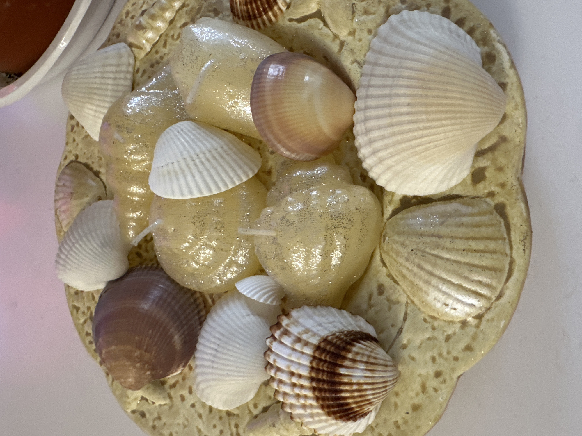 The image shows a decorative object featuring an arrangement of seashells and small, translucent, glittery objects that resemble candies or jewels, set on a beige, porous base. Several types of seashells are present, including small white shells, larger ribbed shells with brown markings, and a smooth, pinkish-brown shell. The beige base is textured with small holes or depressions. The composition is viewed from above. The background is a blurred, white and pink surface. The image does not contain information about Sofia, Bulgaria.