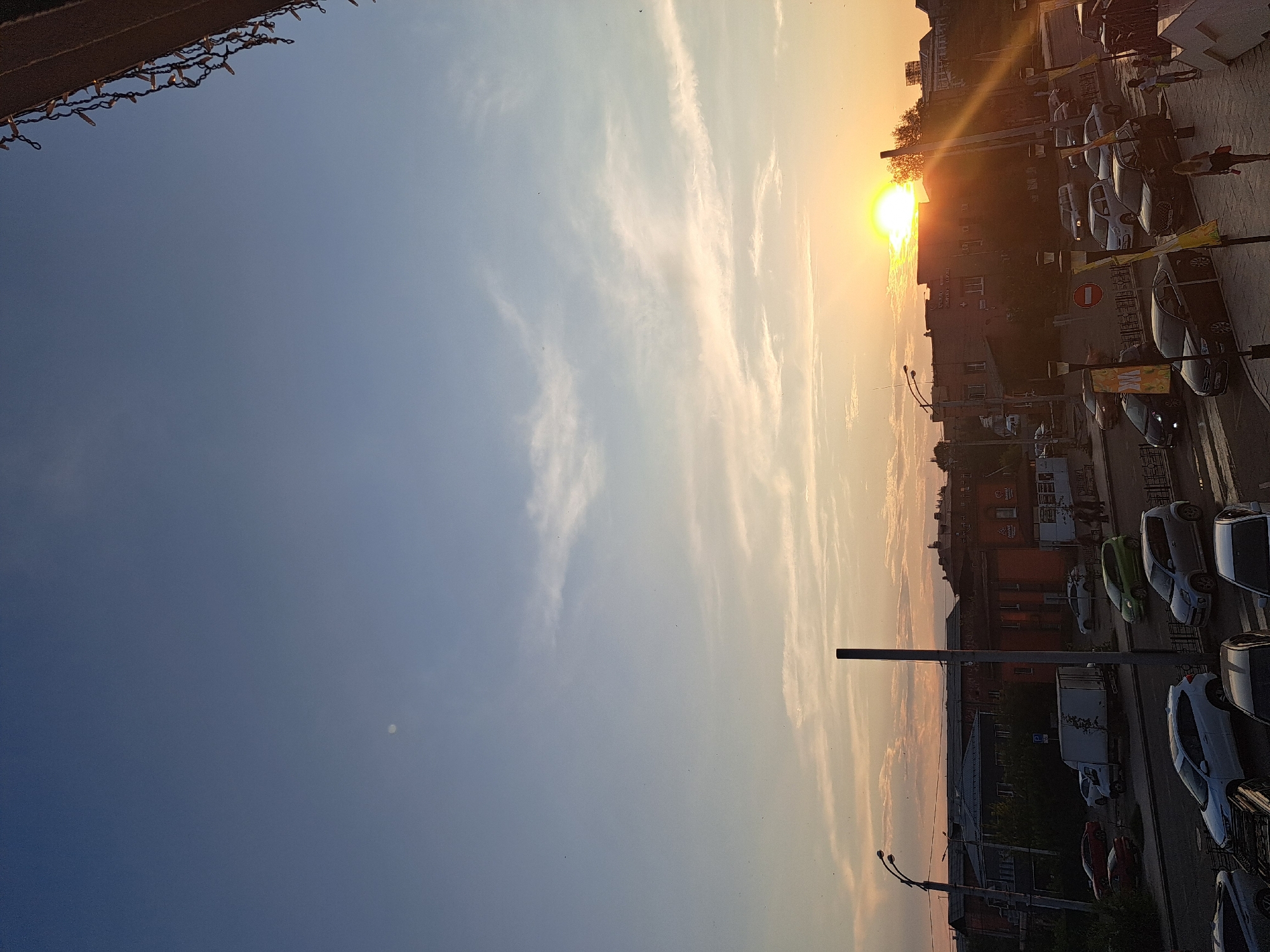 An aerial view captures a sunset over a city street in Irkutsk, Russia, with a clear, pale blue sky transitioning to warmer hues near the horizon. The sun is a bright, white disc peeking from behind buildings, casting a warm glow and long shadows. Several buildings, some appearing residential and others commercial with visible signage, line the street, with varying architectural styles. The street is moderately busy with parked and moving vehicles, including cars, a white truck, and a green hatchback. Pedestrians are visible on the sidewalks. Streetlights and decorative lights are present. The scene appears to be taken from a high vantage point, possibly a balcony or window, overlooking the urban landscape.