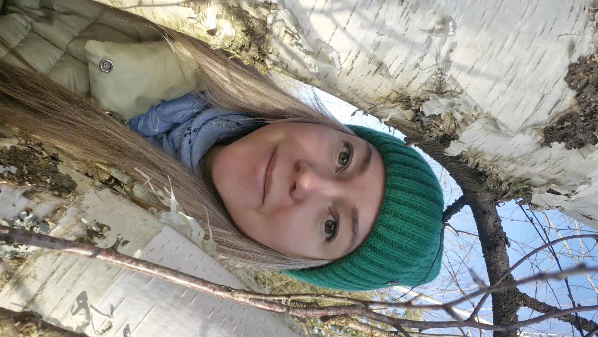 A fair-skinned woman with long blonde hair and a green knitted hat is viewed from a low angle, appearing to be lying under or nestled within a birch tree. She is wearing a light blue scarf and a light-colored quilted jacket. The white bark of the birch tree surrounds her face, with branches visible in the foreground and background. The sky is visible through the branches. The location is Otchor, Russia.