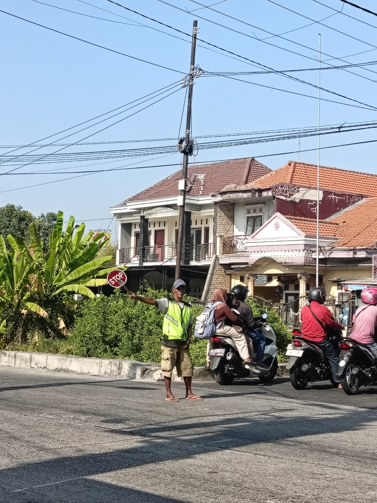 In Gedangan, Indonesia, a man wearing a high-visibility yellow vest and khaki shorts stands in the middle of a street, holding a red and white "STOP" sign. He is directing traffic. Behind him, several motorcycles are stopped. One motorcycle carries a driver and a passenger wearing a hijab and carrying a backpack. Another motorcycle has a driver wearing a red shirt and jeans, and a third motorcycle has a driver wearing a pink shirt and a helmet. The background shows houses with terracotta tile roofs and lush green foliage, including banana trees. Overhead, a complex network of power lines crisscrosses the clear blue sky.